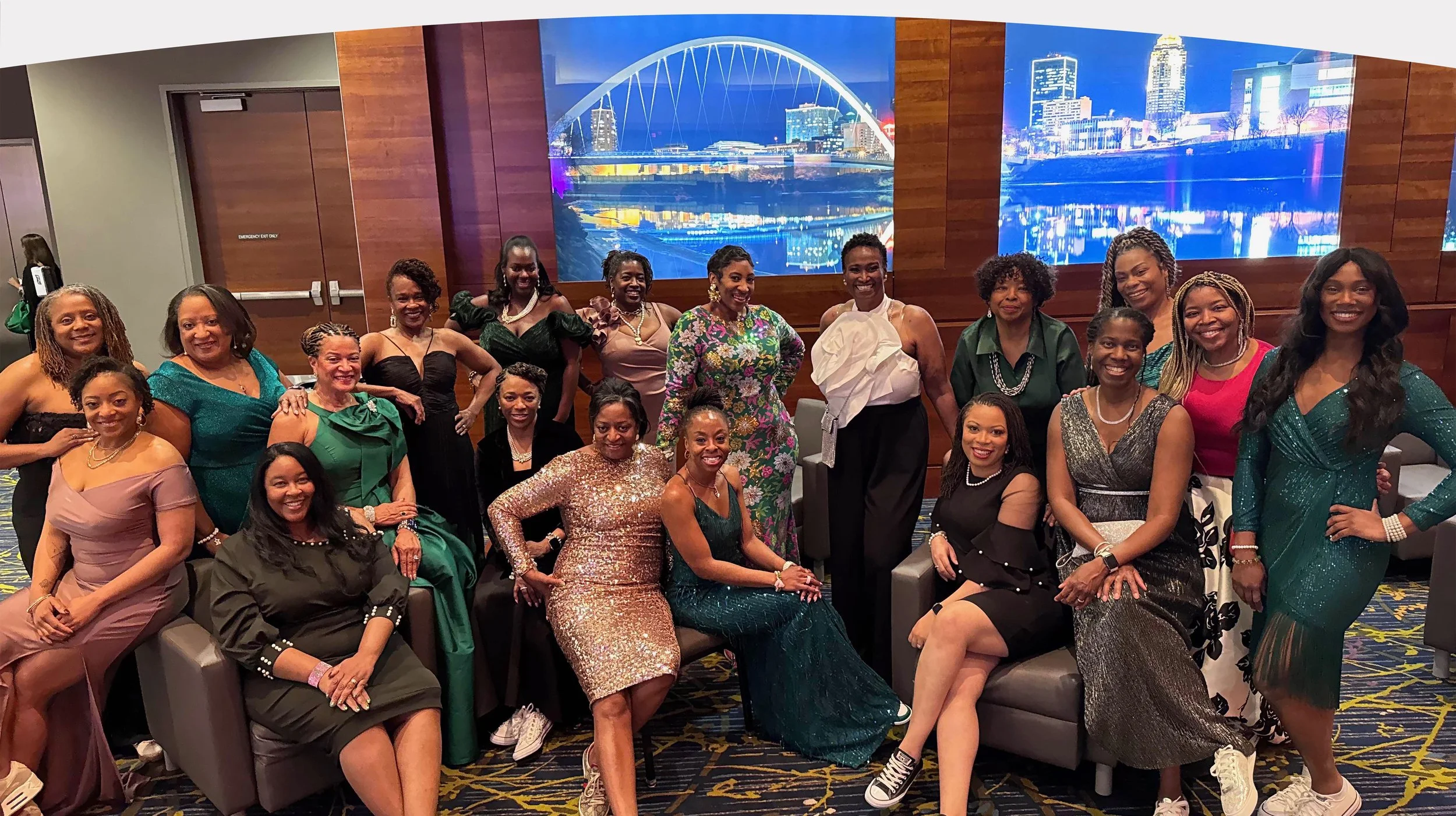 A group of women dressed in formal and colorful attire posing for a photo in an indoor setting with a large digital cityscape display on the wall behind them.