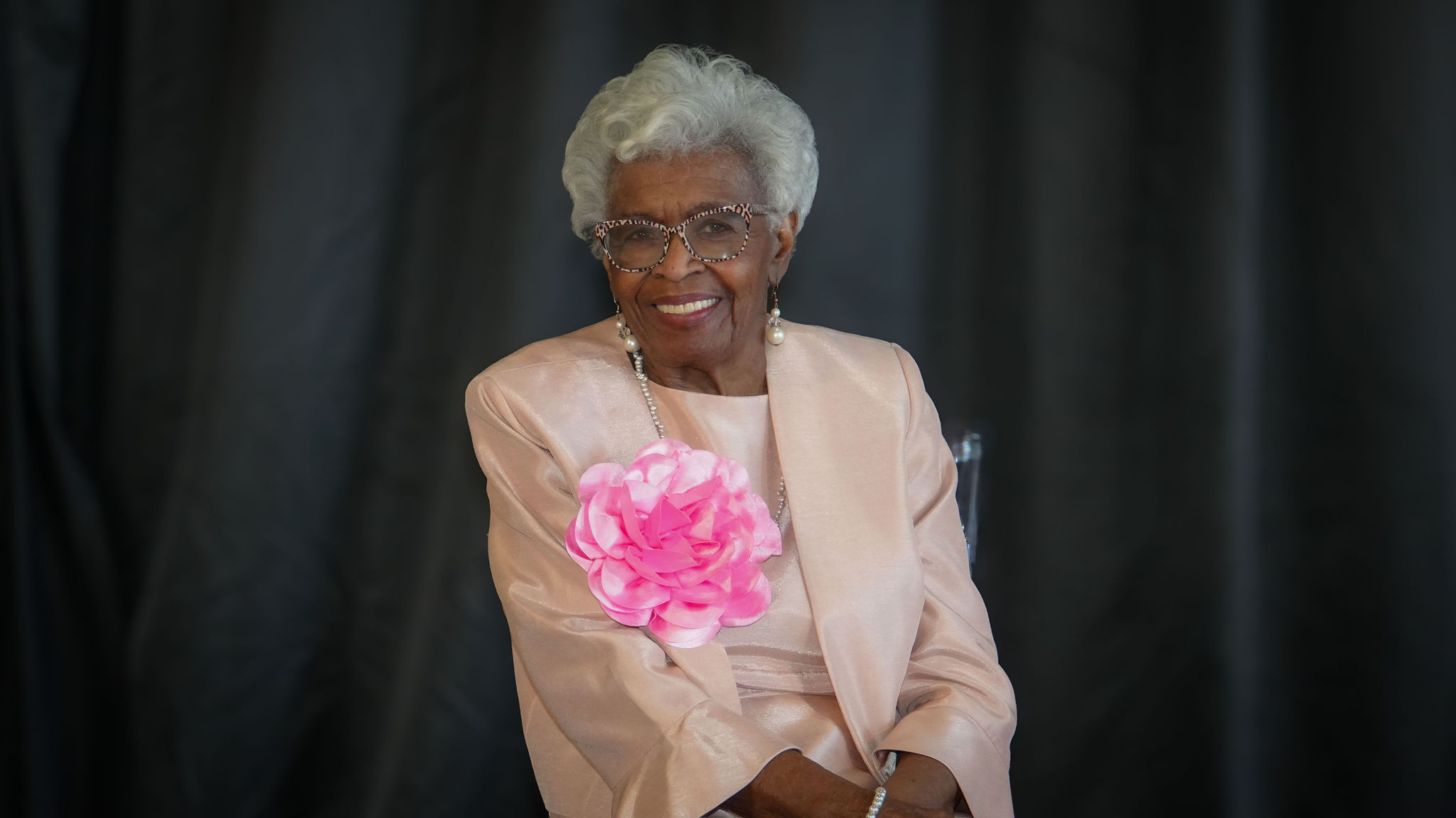 Older woman with short curly gray hair wearing glasses, a light pink suit with a large pink fabric flower on the lapel, pearl earrings, and a pearl bracelet smiling at the camera against a dark background.