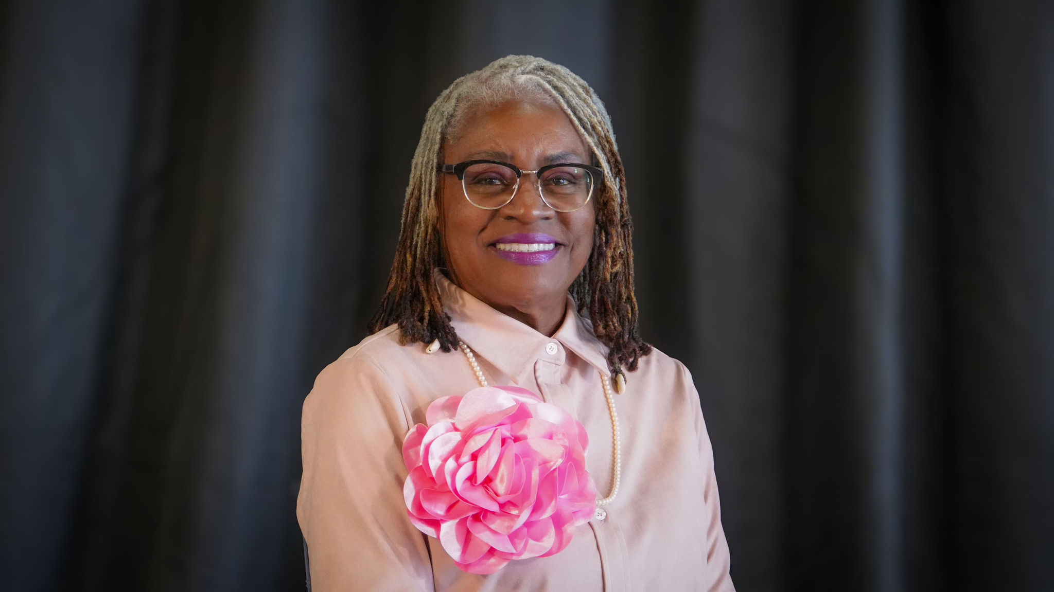 A woman with gray dreadlocks and glasses smiling, wearing a pink blouse with a large pink floral attachment and a pearl necklace, in front of a dark curtain.