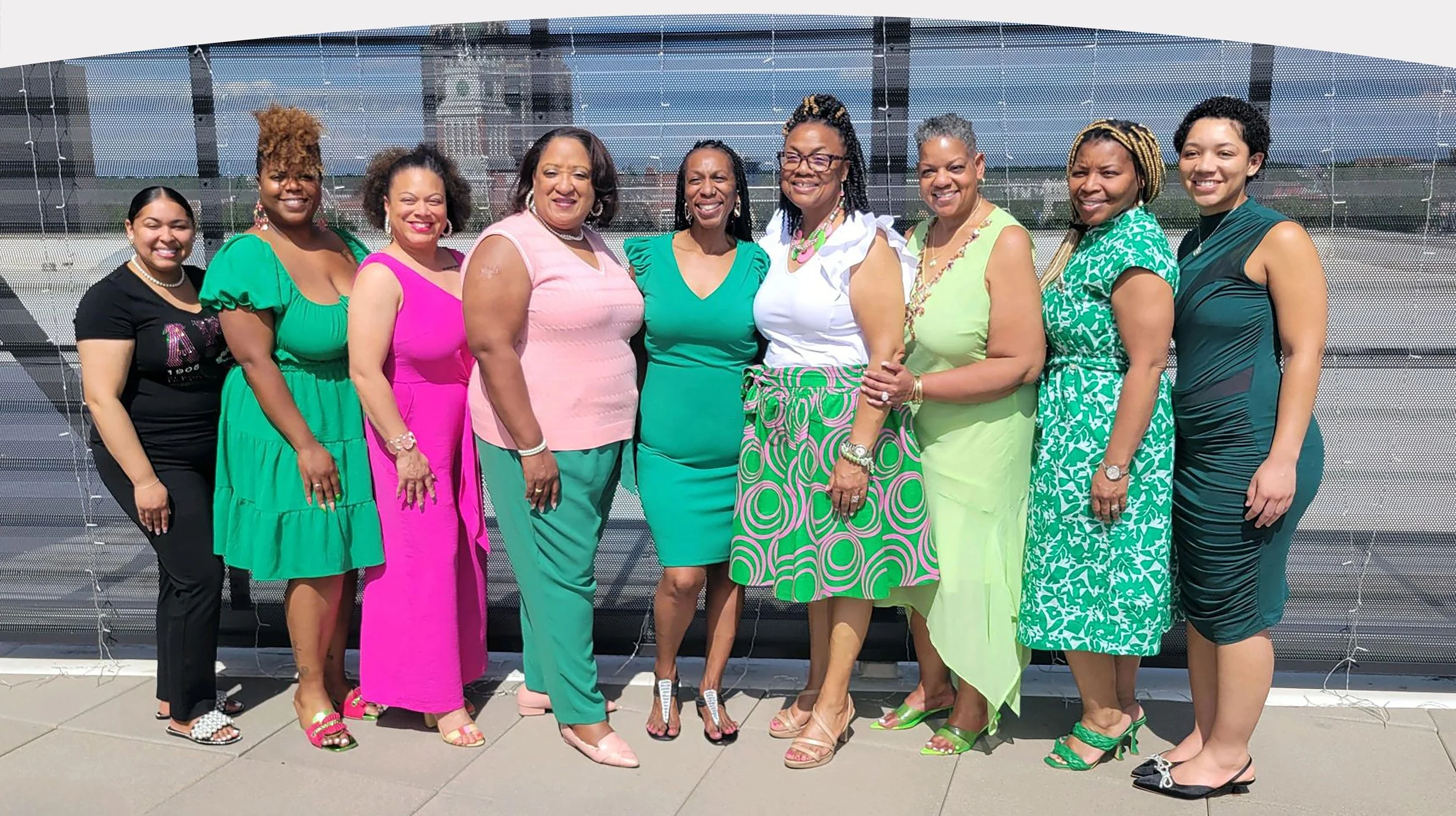 A group of eleven women standing together outdoors, dressed in colorful summer attire, in front of a modern building with a glass facade.