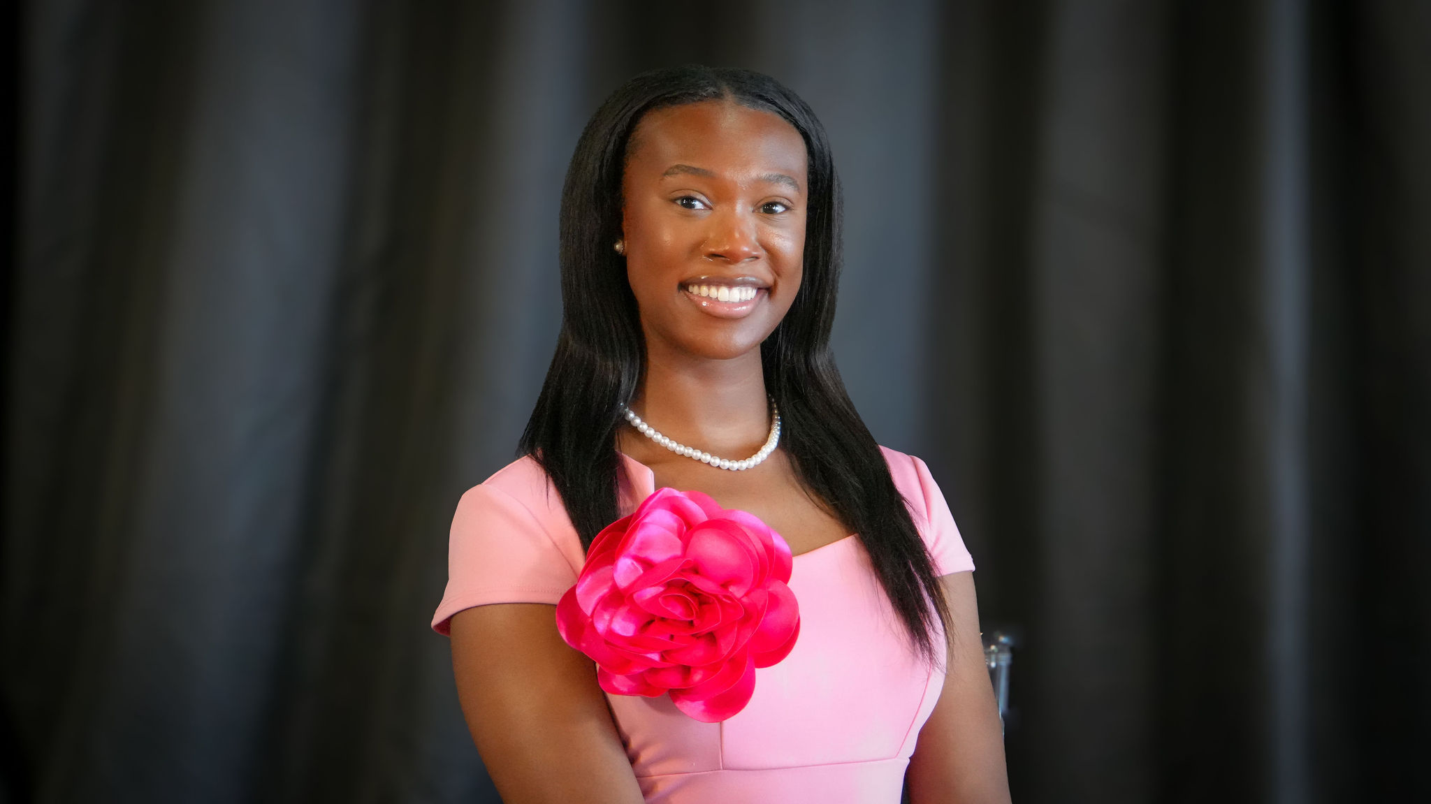 A young woman smiling, wearing a pink dress with a large pink fabric flower on the shoulder, a pearl necklace, and pearl earrings, standing against a blurred dark background.