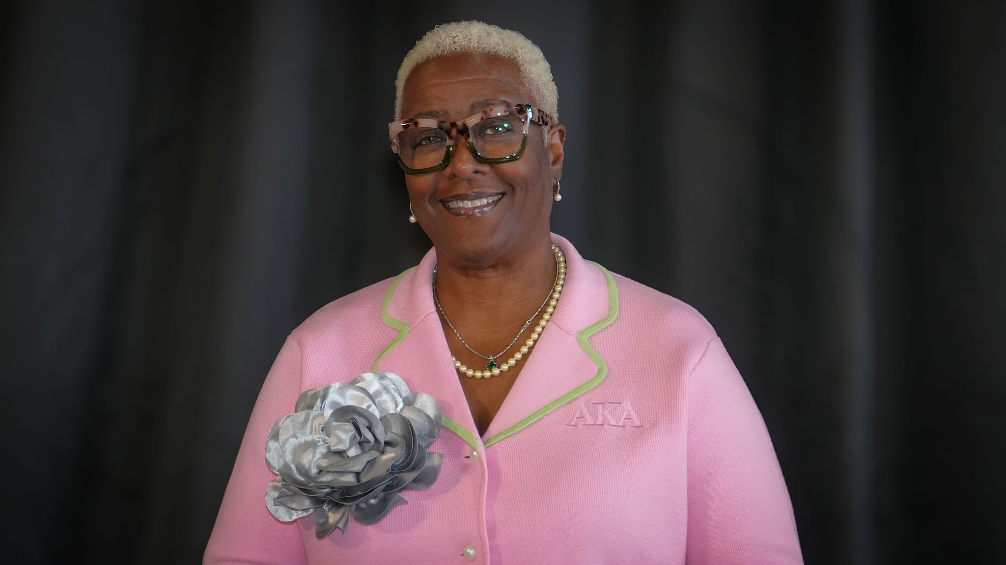 An older woman with short, bleached blonde hair and glasses, wearing a pink blazer with green trim, pearl earrings, and a pearl necklace, smiling against a dark background.