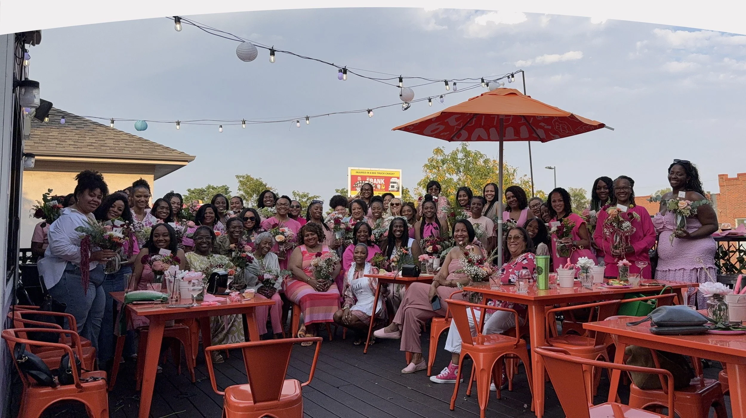 A large outdoor gathering of women, many in pink outfits, holding bouquets, sitting and standing around tables decorated with flowers, under string lights and an orange umbrella, on a patio with sky and trees in the background.