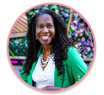 A woman with long curly black hair smiling, wearing a green blazer and a white blouse with a pearl necklace, standing outdoors with colorful flowers and greenery in the background.