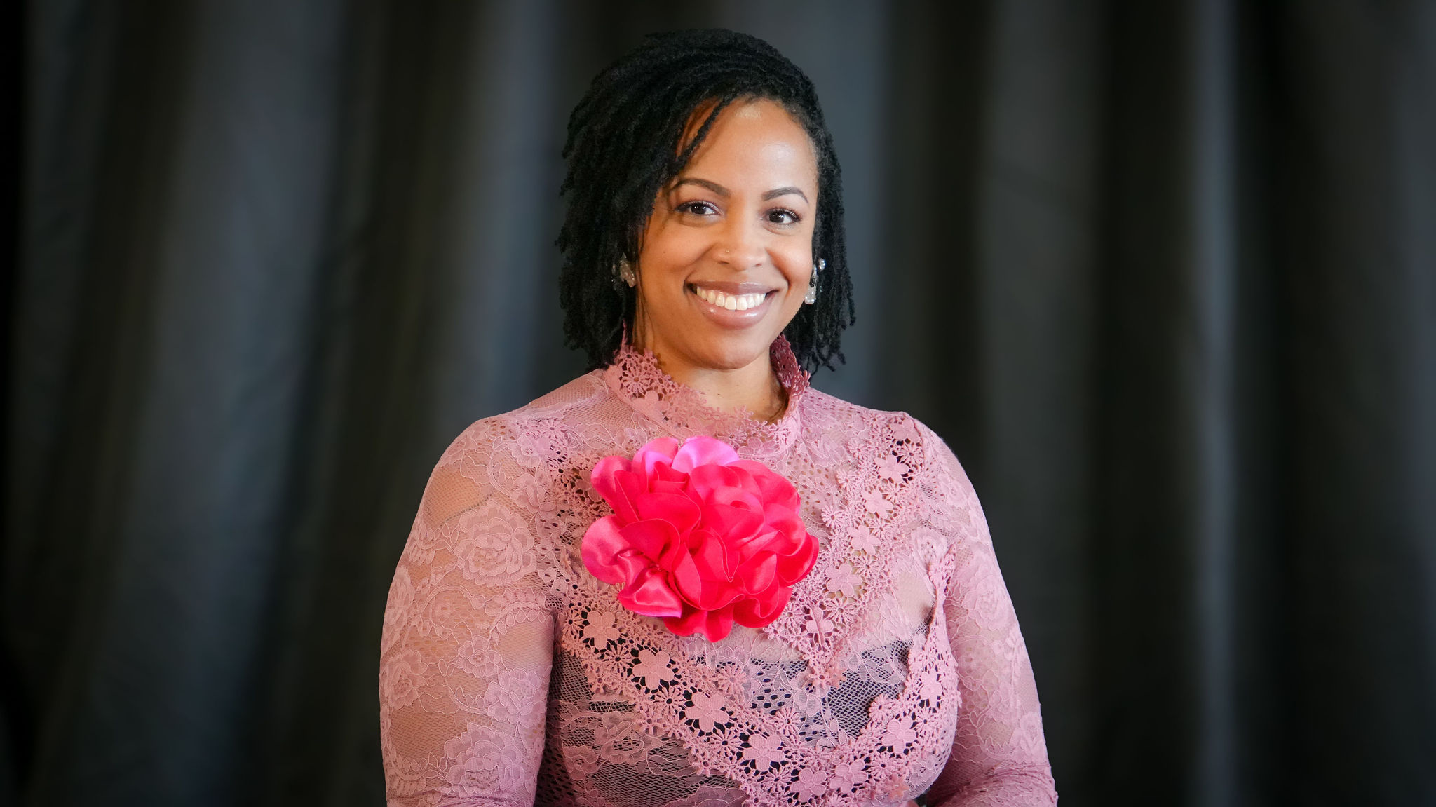 A woman smiling, wearing a pink lace dress with a large pink floral corsage on her chest, earrings, and sporting dreadlocks, standing against a dark background.