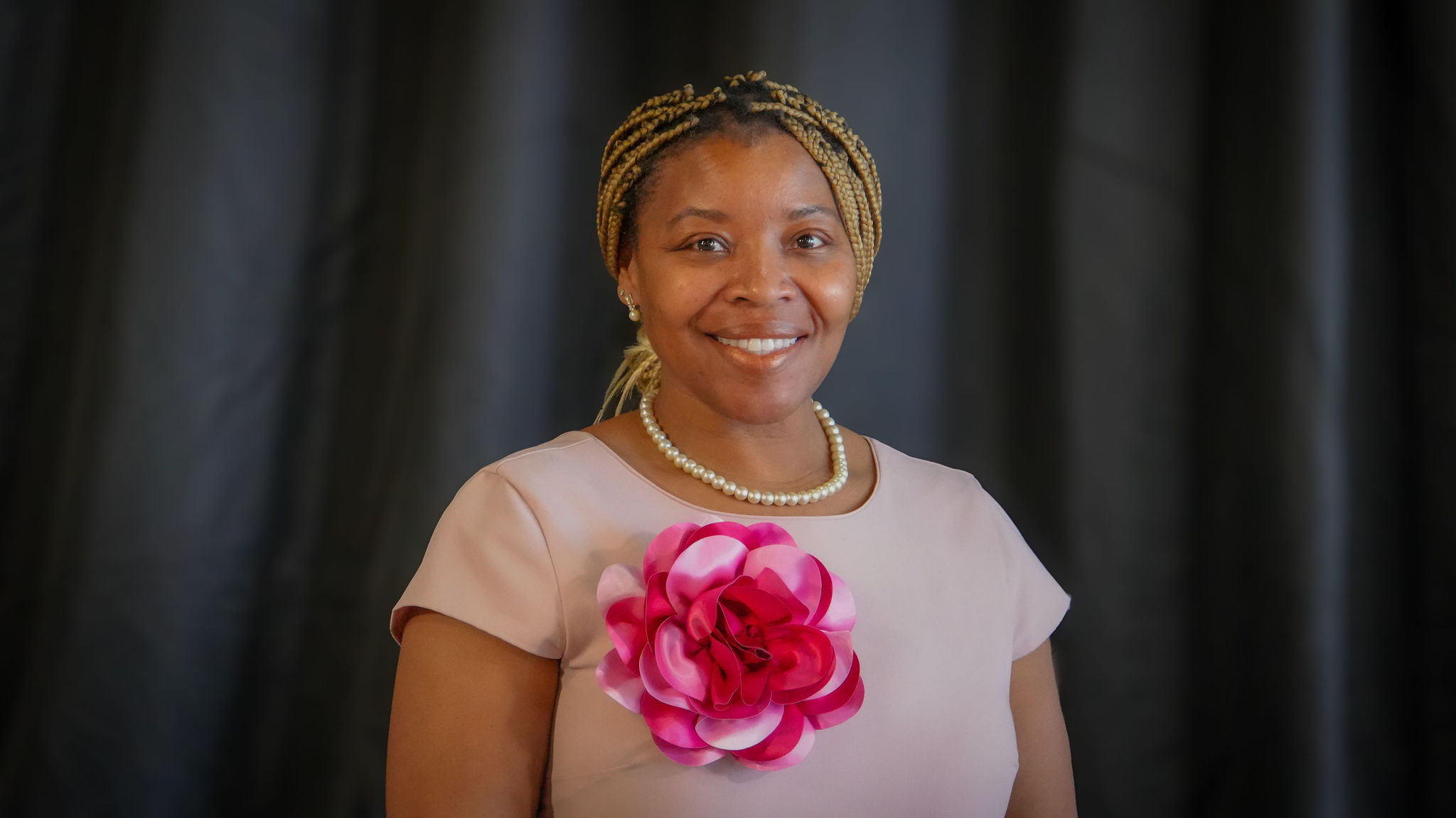 A woman wearing a pearl necklace and earrings standing in front of a dark background, smiling, with a large pink and white floral decoration on her dress.