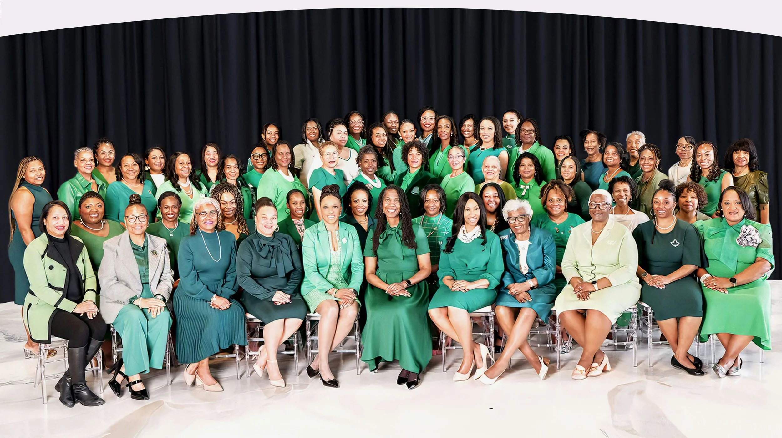 Group photo of diverse women dressed in green, sitting and standing on a stage with a black curtain background.