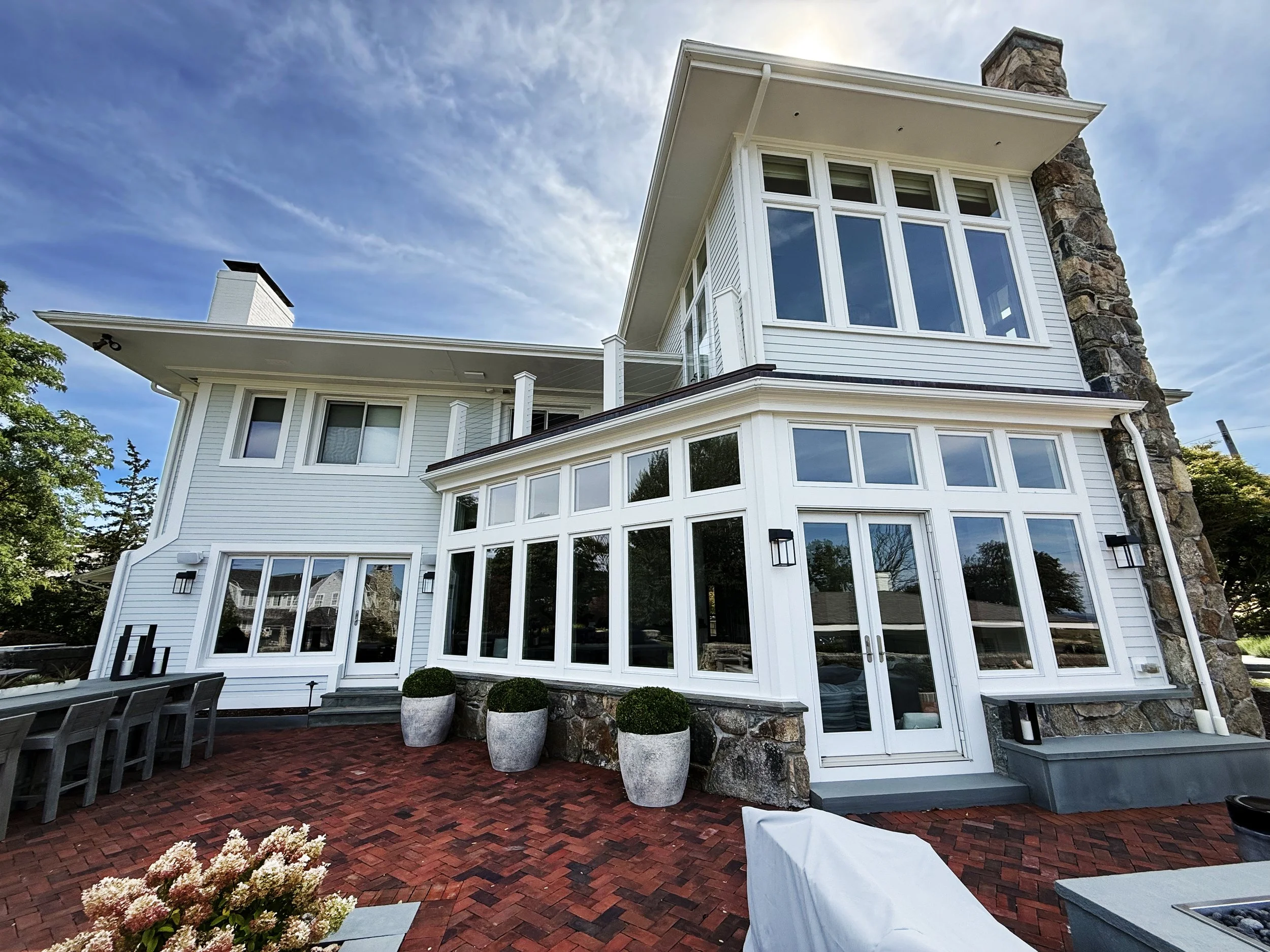 Exterior view of a modern multi-story house with white siding, large windows, and a stone chimney. The house features a brick patio with outdoor furniture and potted plants, under a partly cloudy sky.