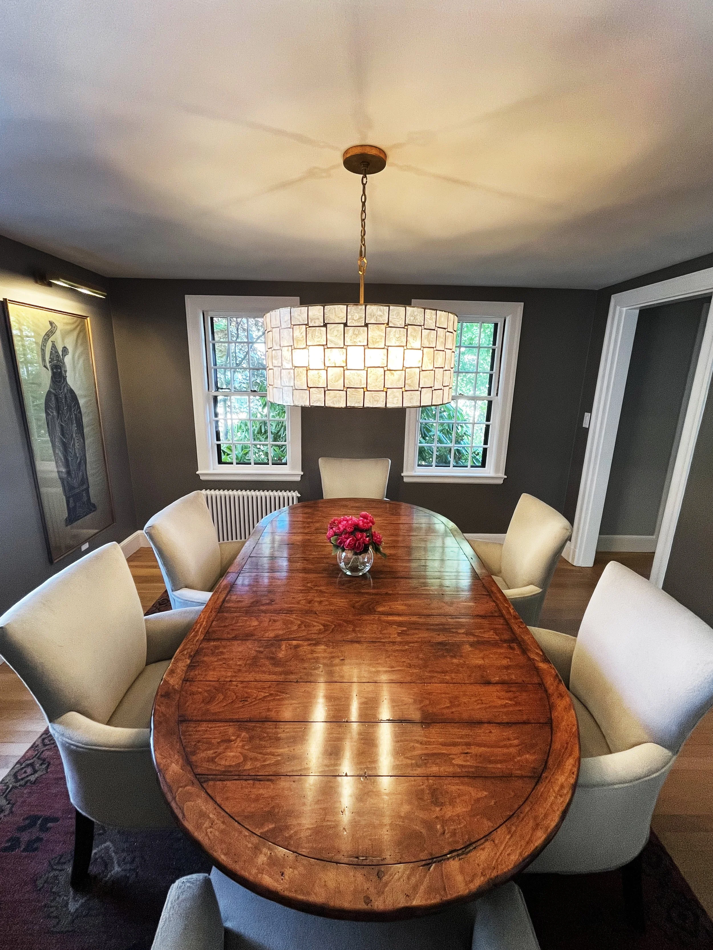 A cozy dining room with a wooden oval table and six cushioned beige chairs. A clear vase with pink flowers is centered on the table. A large, circular, tiled chandelier hangs above. The room has dark gray walls, white window frames, and two windows with greenery outside. There's a portrait on the left wall and a radiator below the windows.