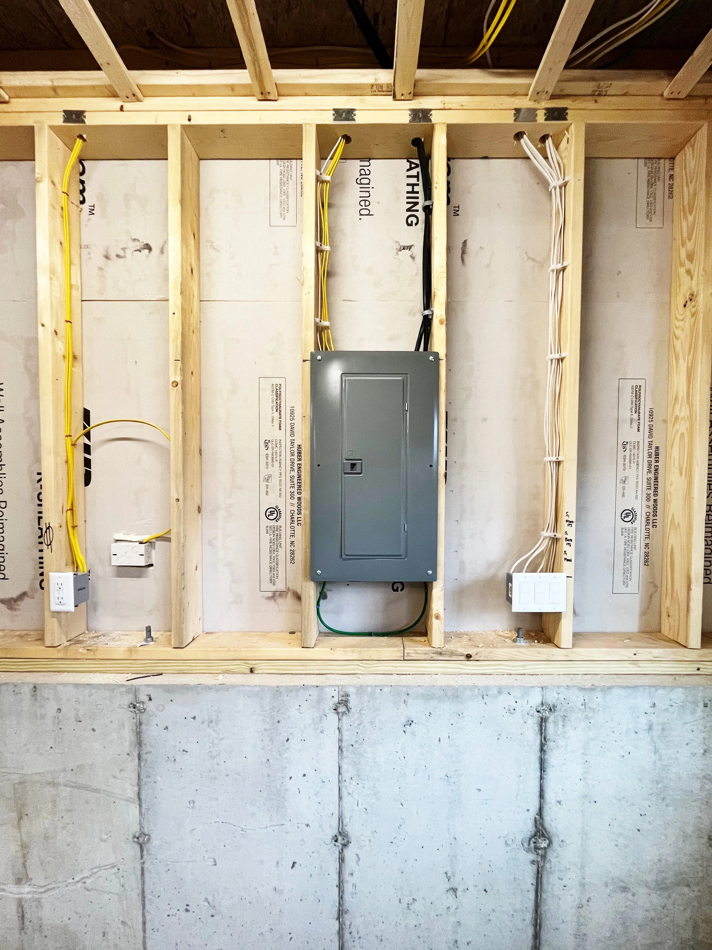 Electrical wiring and a grey electrical panel mounted in an unfinished wall frame, with yellow, black, and white wires connected.