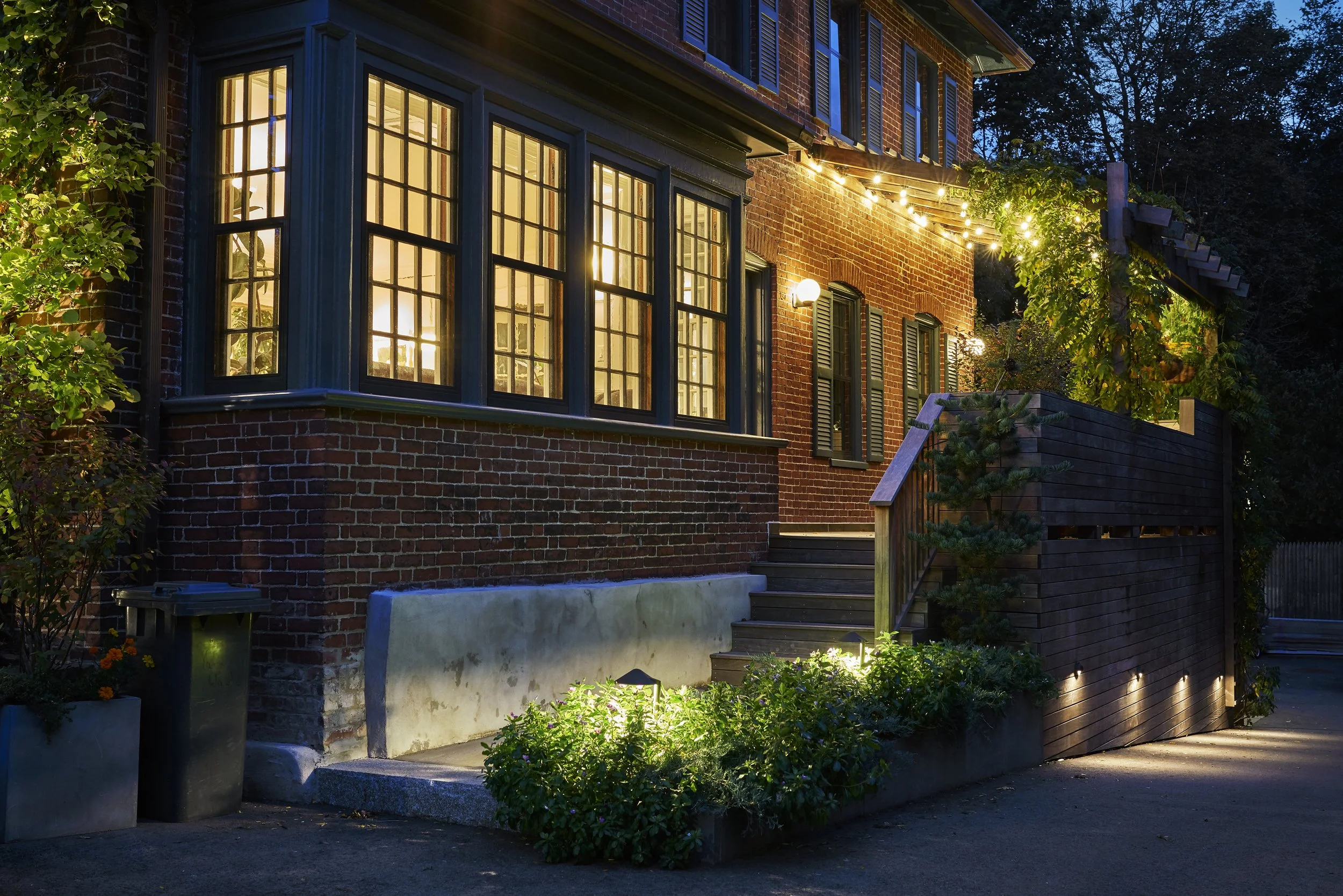 A cozy brick house at night with warm interior lights, string lights outside, and a small garden with lighting along the walkway.