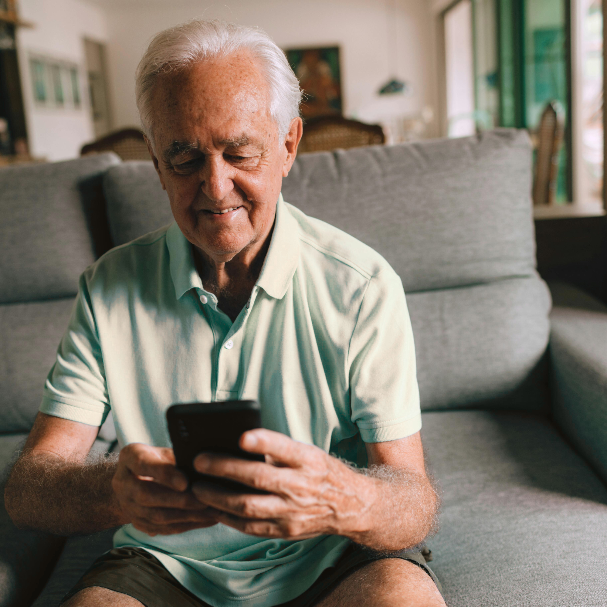 Older man sitting on a couch, smiling and looking at a smartphone in his hands, in a well-lit living room.