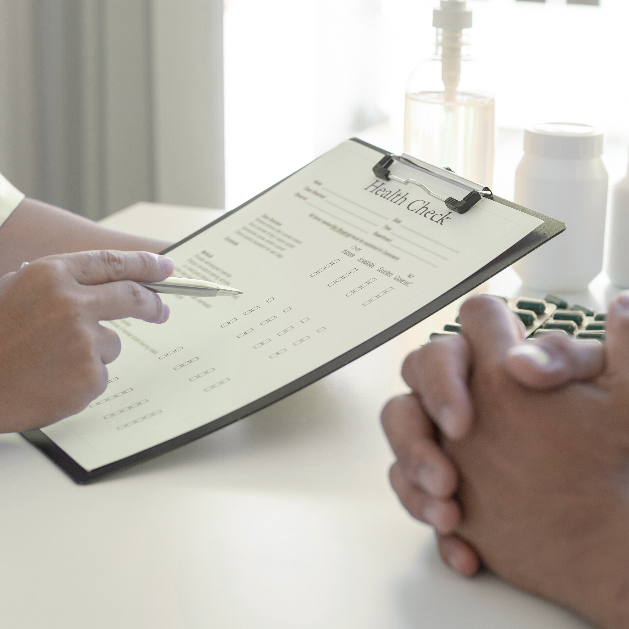 A healthcare professional holding a pen and reviewing a health check form on a clipboard, with a patient's hands clasped on the table.