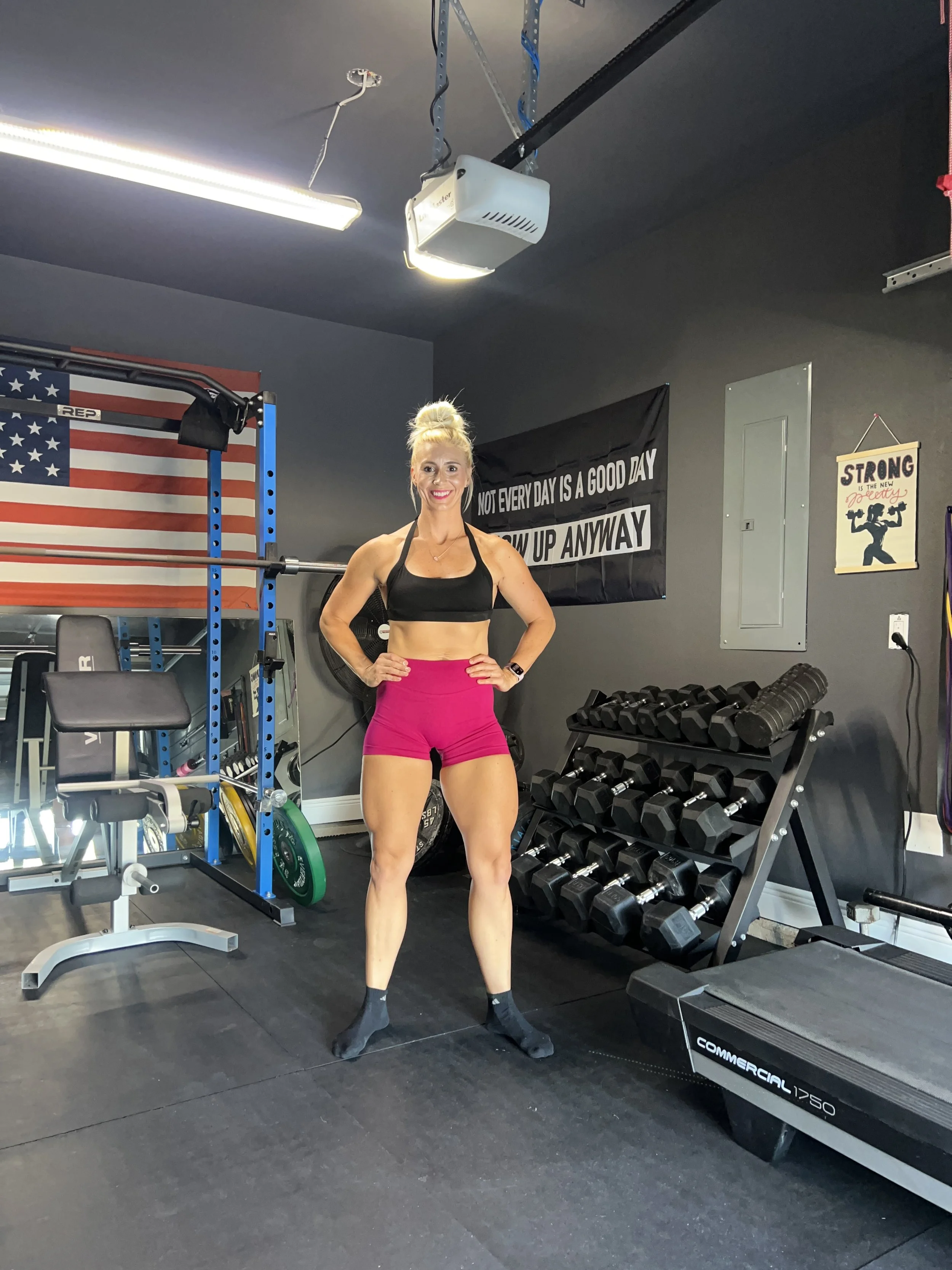 A woman in black sports bra and pink shorts standing in a home gym with hands on hips, smiling at the camera.