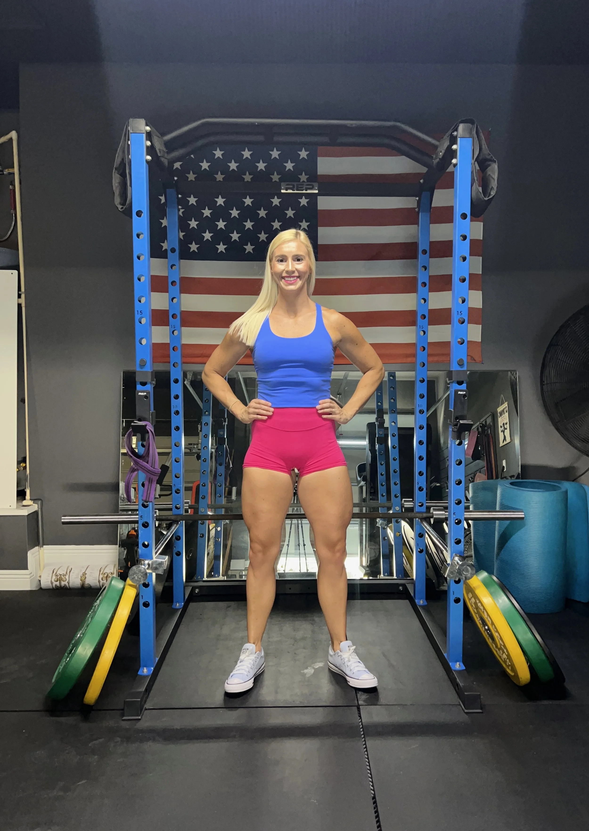 A woman in a blue tank top and pink shorts standing in a gym in front of a barbell and weight plates, with an American flag hanging on the wall behind her.