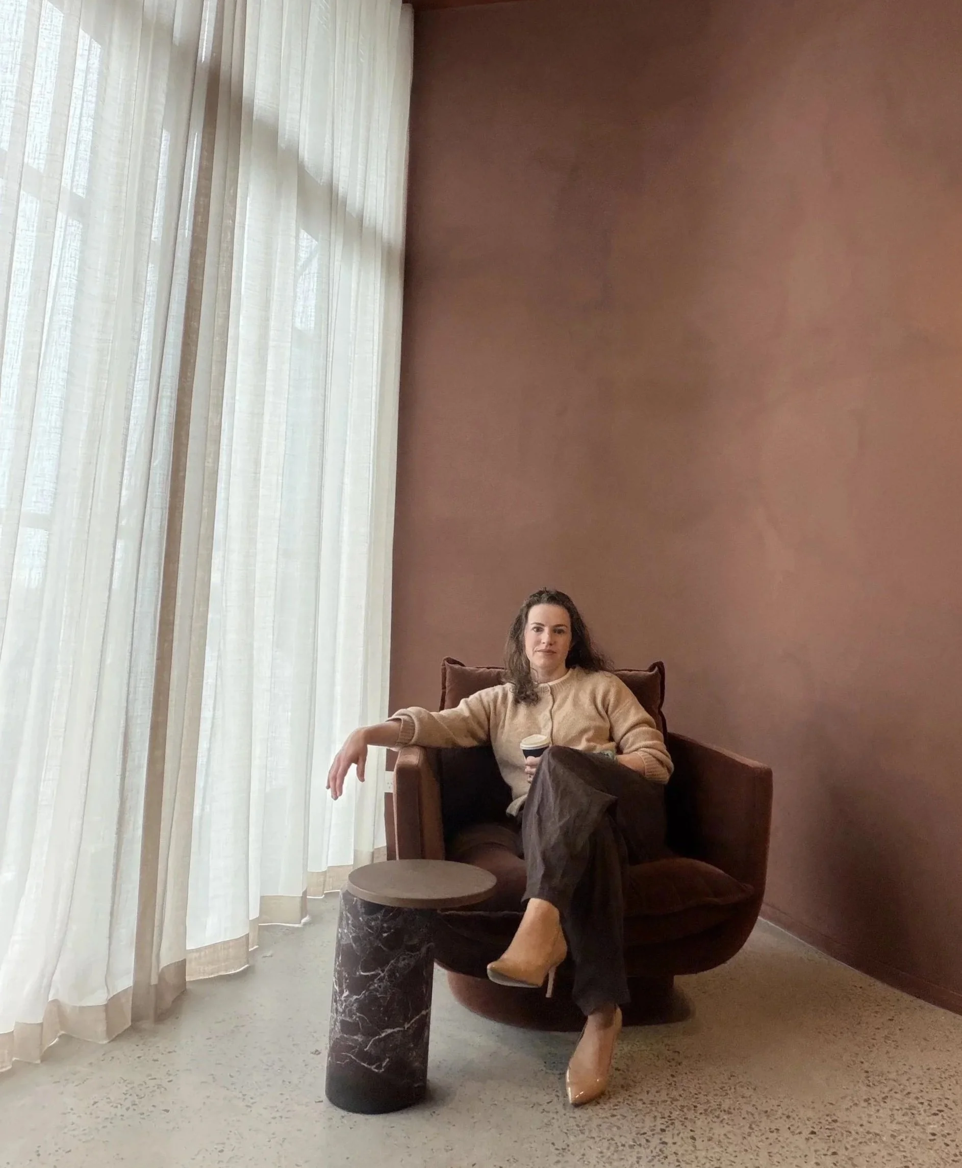 Dr Hannah Dobson sitting in a brown armchair in a room with large window curtains, holding a coffee cup, wearing a beige sweater, black pants, and beige pointed-toe shoes, with a small round marble side table next to her.