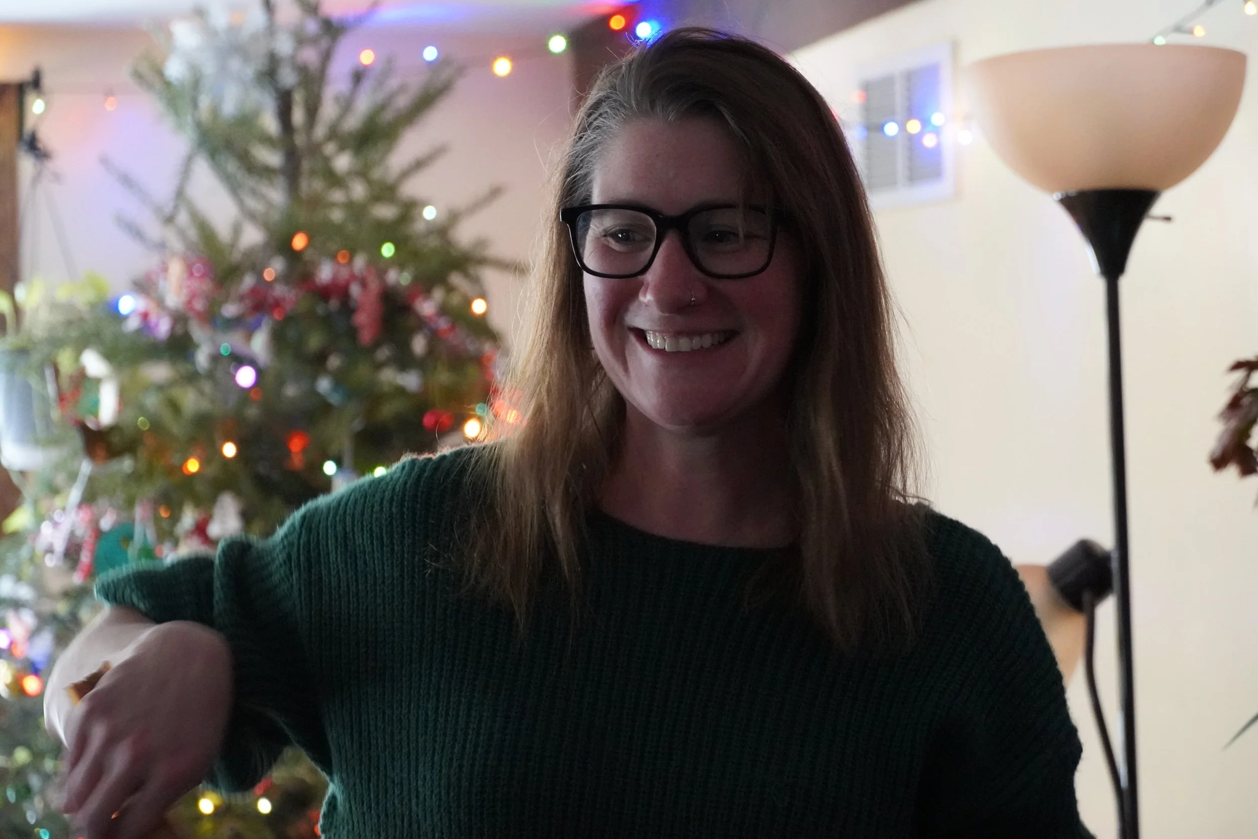 A woman with glasses smiling, standing near a decorated Christmas tree with lights in a cozy indoor setting.