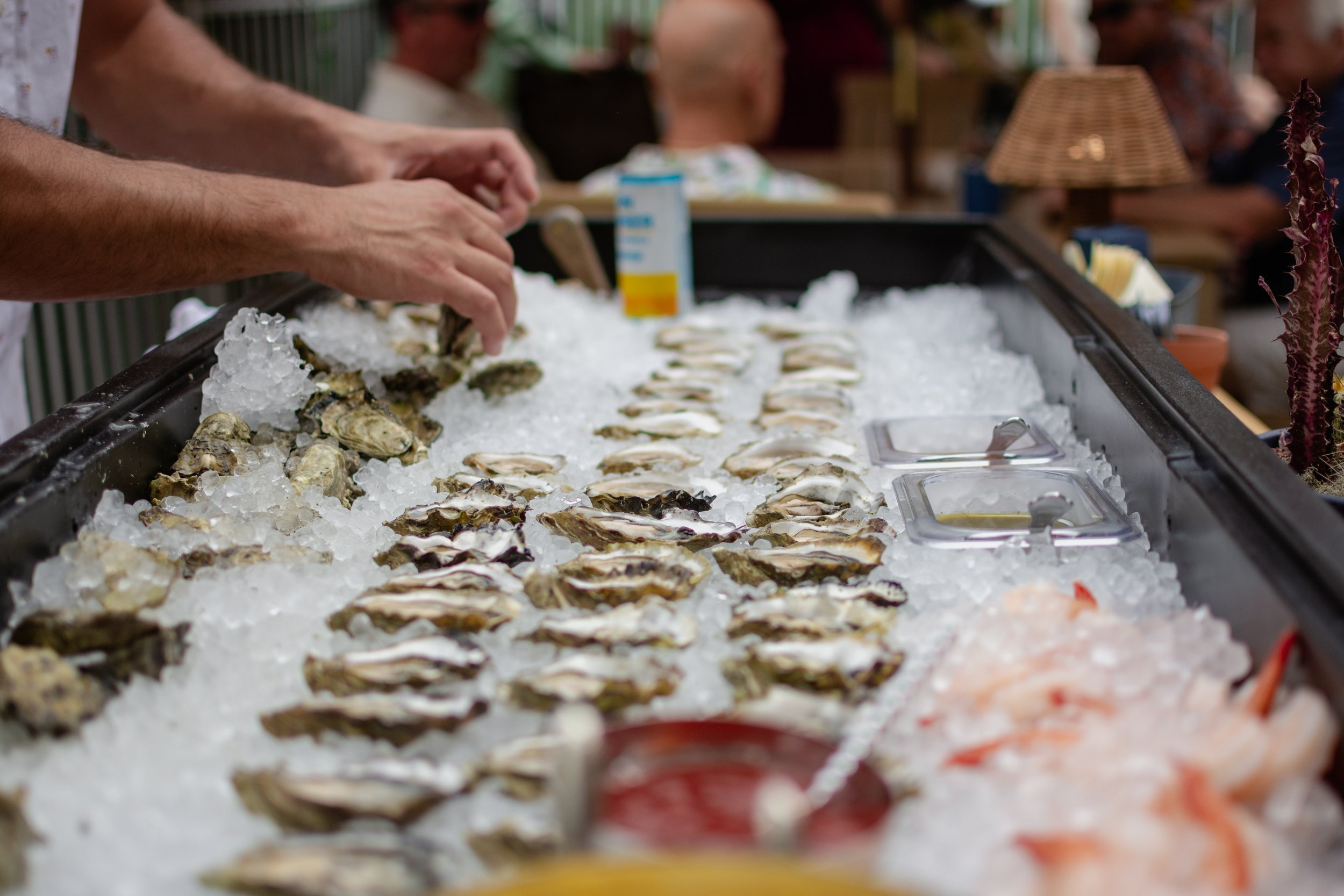 A person shucking oysters on ice at a seafood market or restaurant, with other customers in the background.