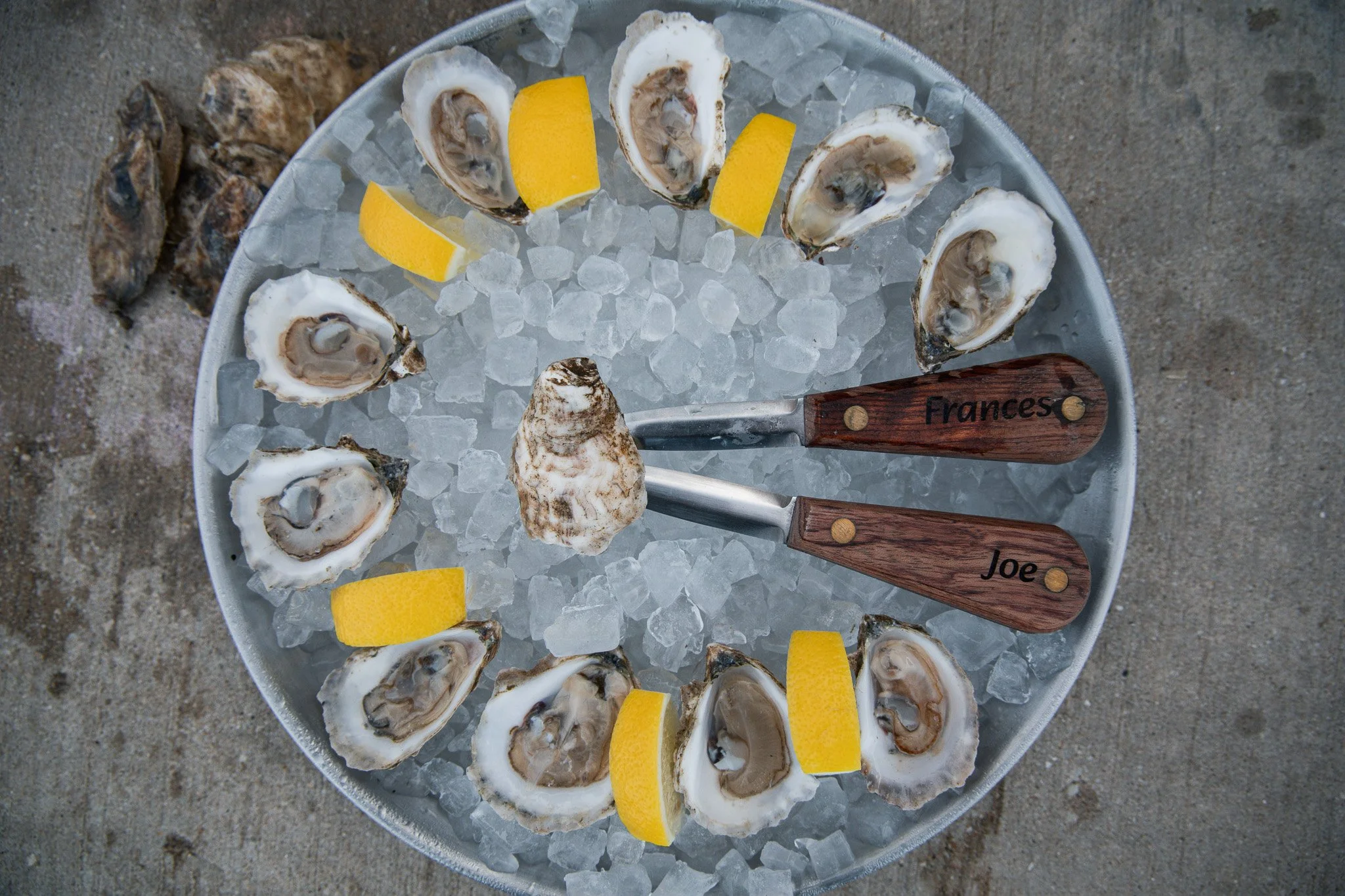 A round serving tray filled with ice, oysters on the half shell, and lemon wedges. Four wooden-handled knives, engraved with names 'Frances' and 'Joe,' are resting on the tray. The tray is placed on sand, suggesting an outdoor setting.