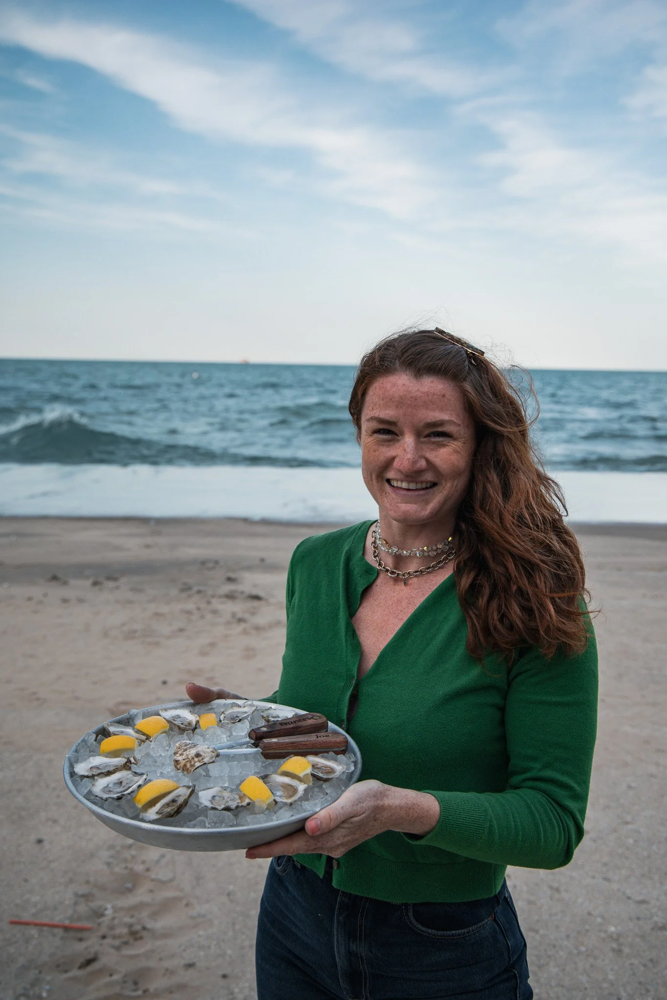 A woman with long, curly red hair, wearing a green cardigan and jewelry, smiling while holding a tray of oysters and lemon wedges on the beach with the ocean and sky in the background.