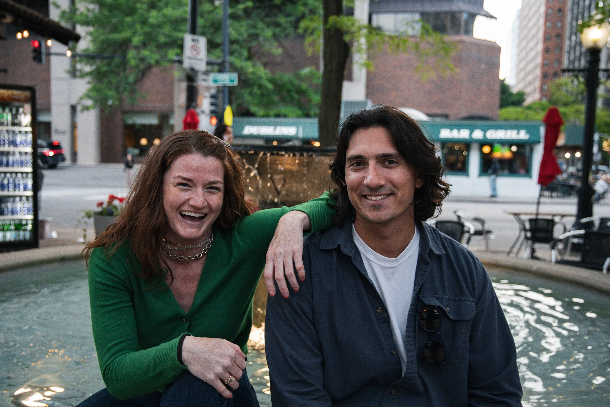 A woman with red hair and a man with dark hair smiling and sitting by a fountain in an urban park, with a bar and grill, trees, and buildings in the background.