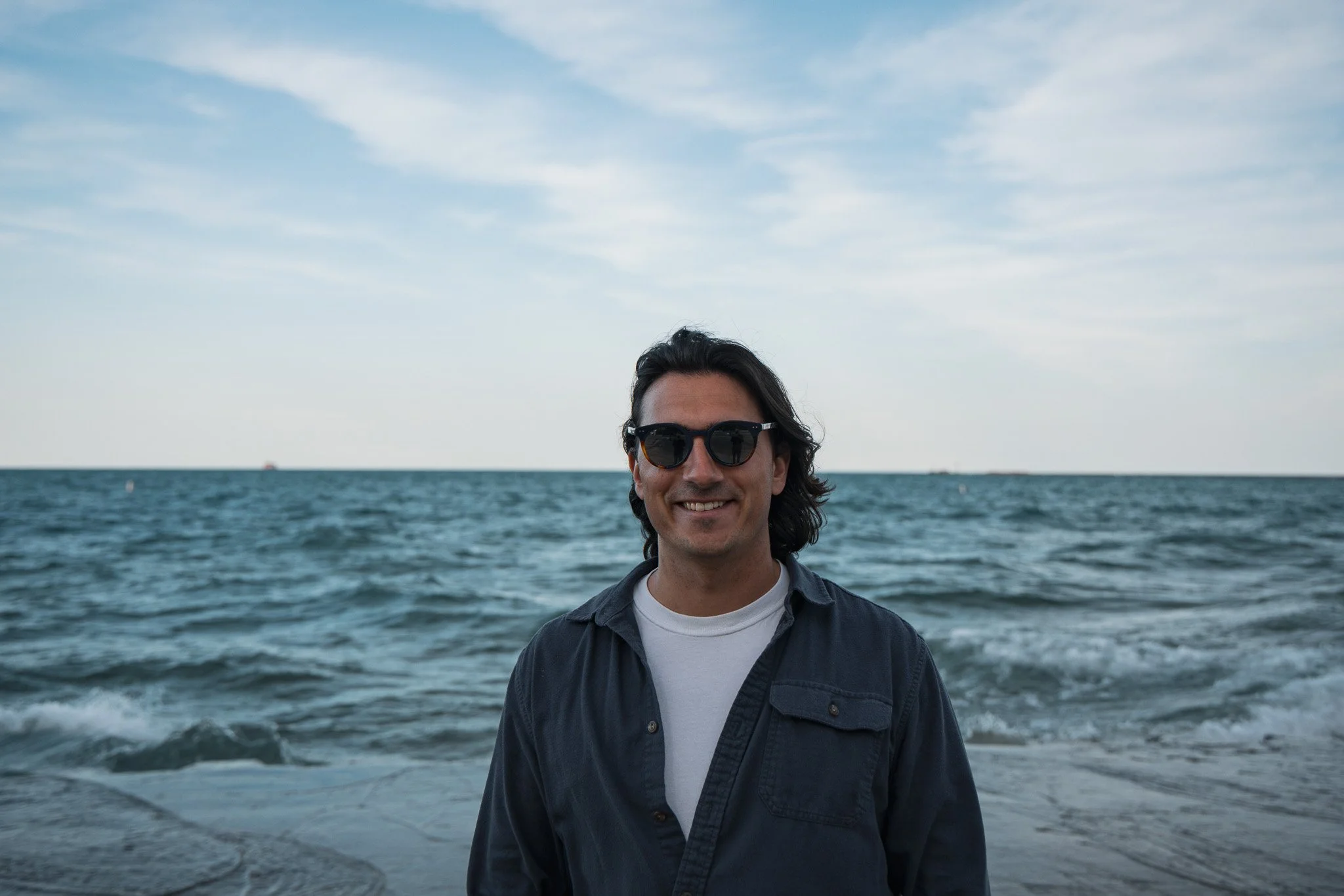 A man with dark hair, wearing sunglasses, a white t-shirt, and a dark shirt, smiling while standing on a boat or near water, with a cloudy sky and open water in the background.