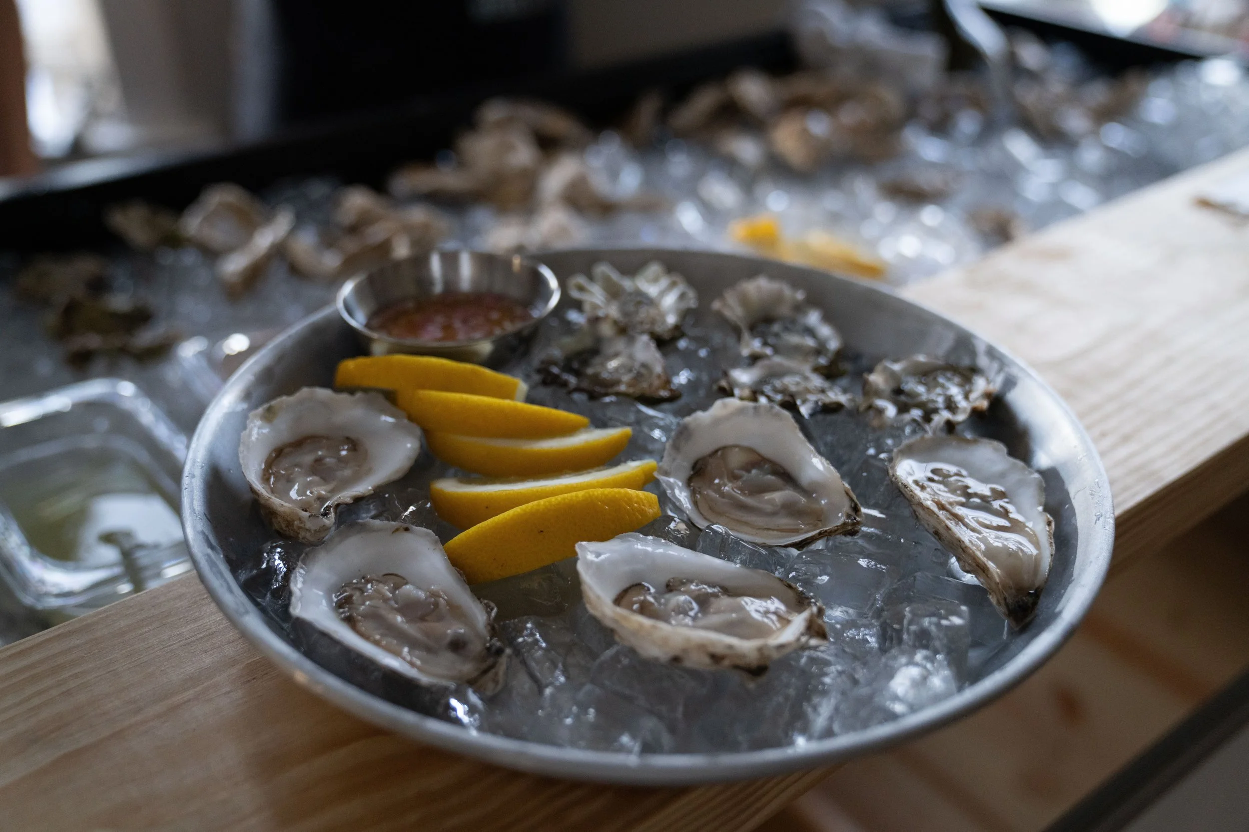 A tray of opened oysters on ice garnished with lemon wedges and a small cup of cocktail sauce. In the background, more oysters are visible on a bed of ice.