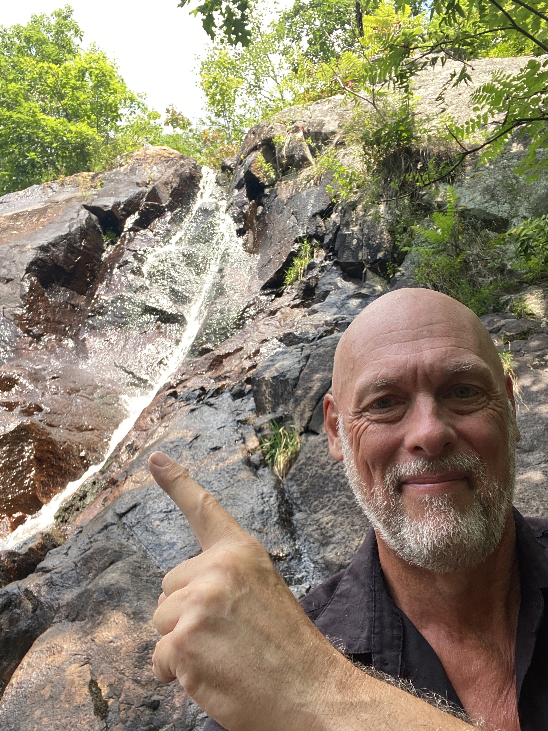 An elder with a beard smiling and pointing at a waterfall in a forested area.