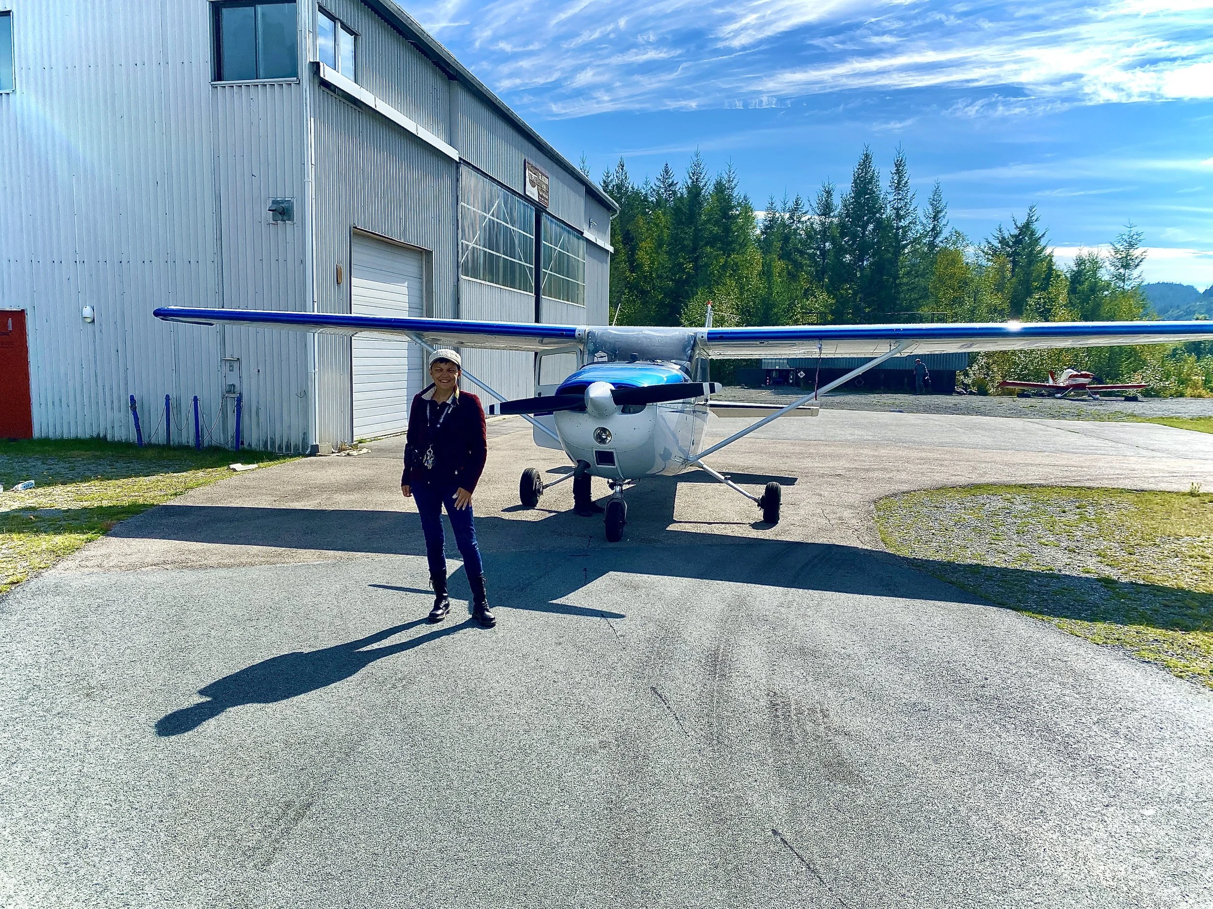 A woman standing in front of a small aircraft parked outside a metal hangar. The woman is wearing a cap and jacket, smiling at the camera. The background features a clear blue sky, some trees, and another small plane parked further away.