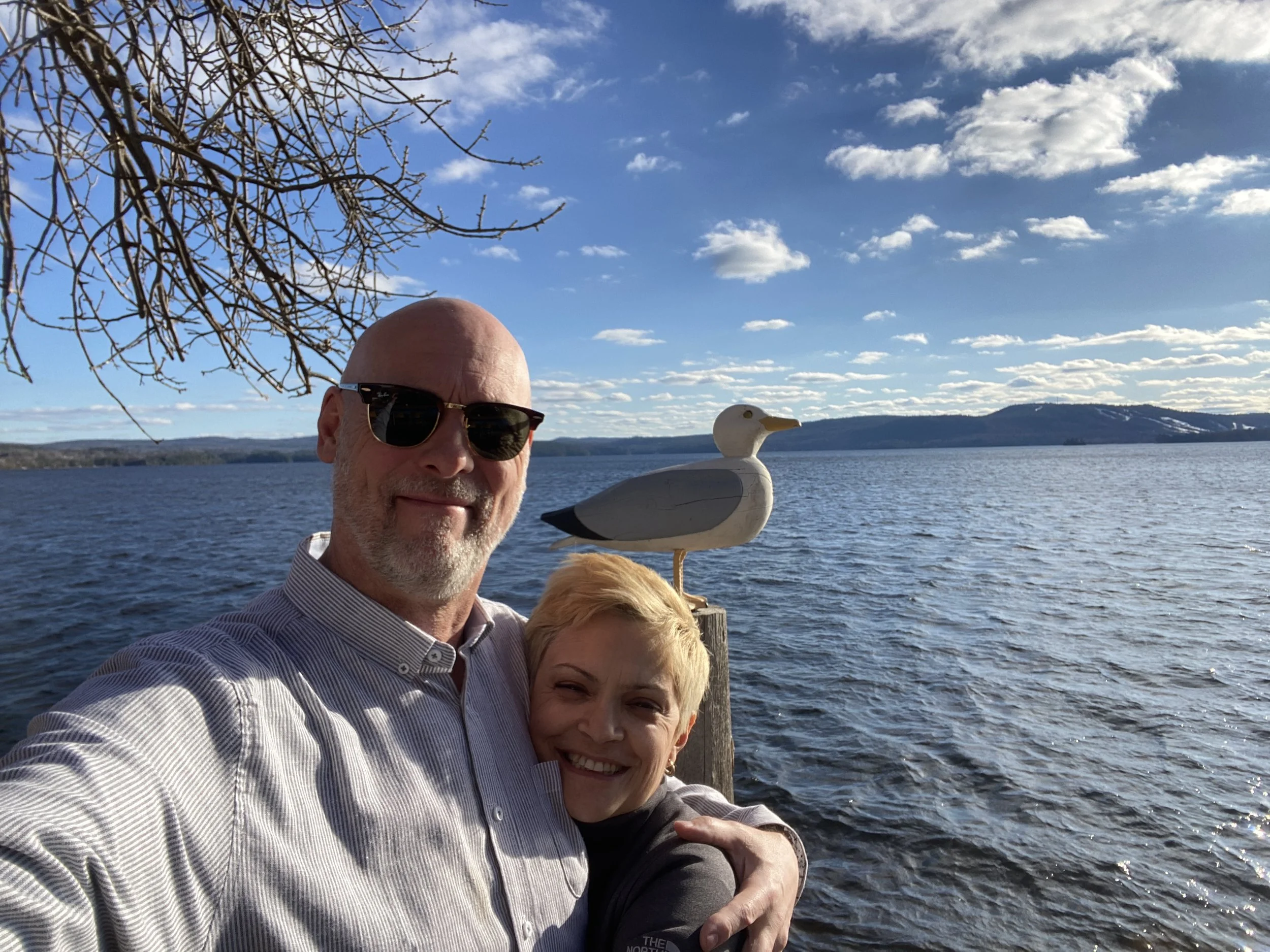 A man with sunglasses and a woman smiling, standing near a body of water, with a dog in front of them. A duck-shaped sculpture or decoration is on a post beside the water, and a scenic landscape with hills and a partly cloudy sky is in the background.