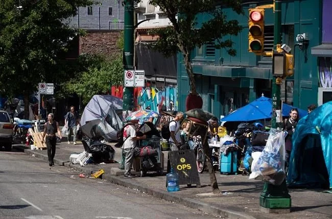 Street scene in an urban area with people, tents, and umbrellas set up for a street market or homeless encampment. Several pedestrians walk along the sidewalk, and a traffic light is visible.