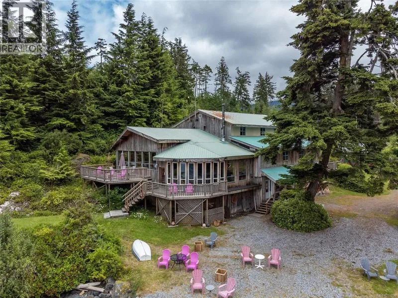 Large wooden house with a green metal roof is surrounded by trees in a lush forest, with outdoor chairs and tables in the yard.