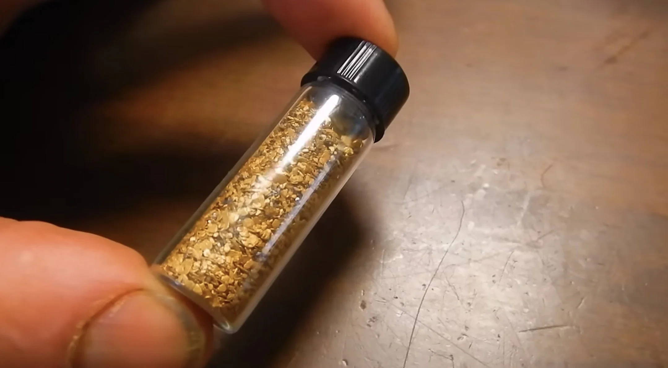 A hand holding a small glass vial containing golden-colored crushed material with a black screw-on lid.