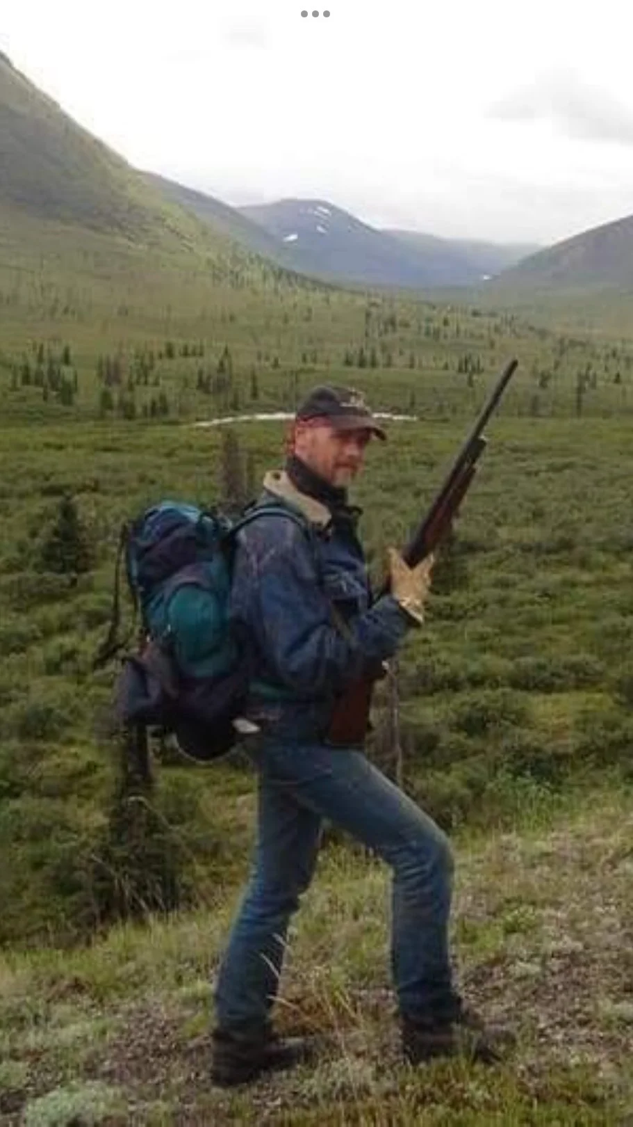 A man in outdoor gear holding a rifle in a green mountainous landscape.