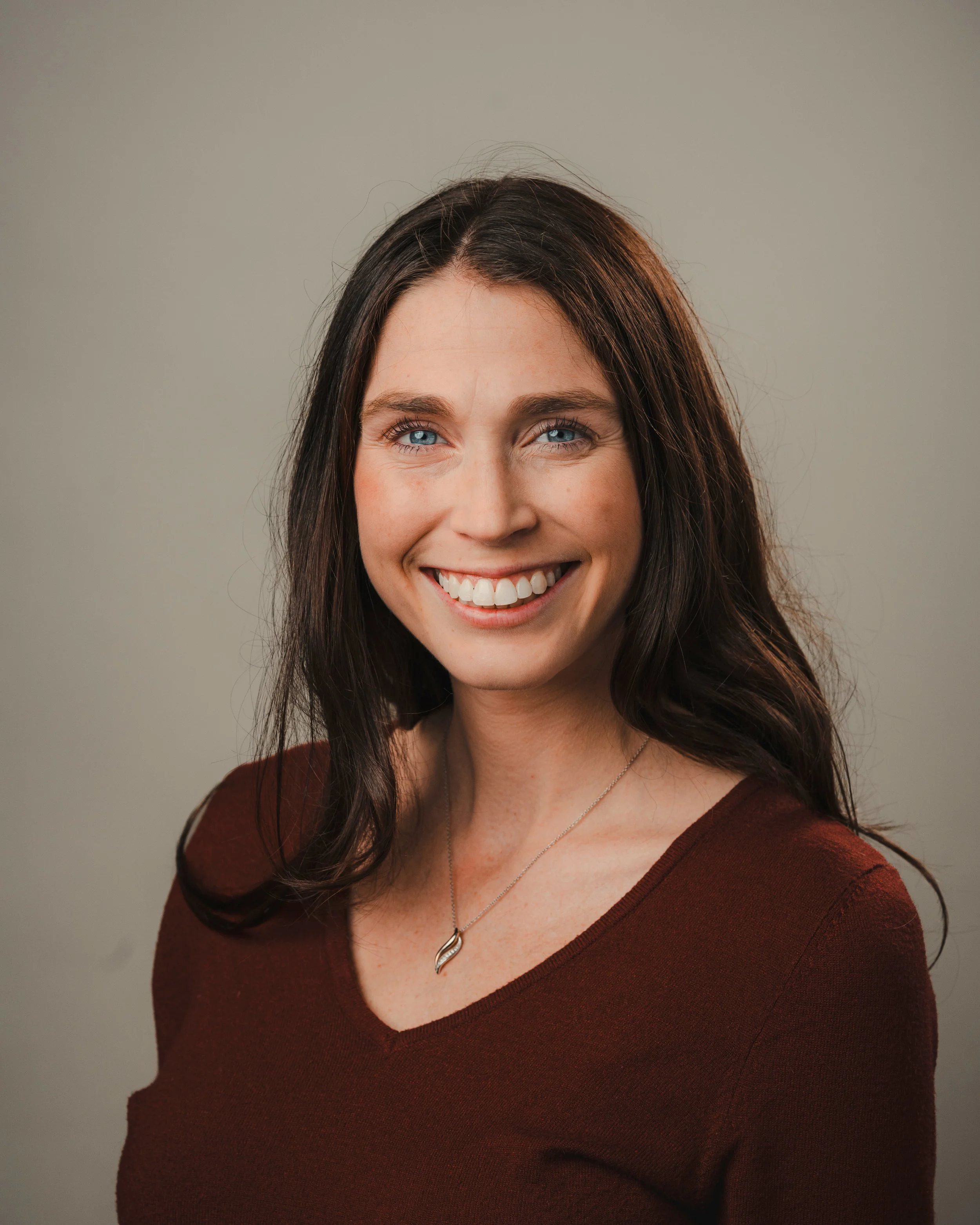 Kerry Maska. A smiling woman with blue eyes, wavy dark brown hair, wearing a maroon top and a silver necklace, against a neutral background.