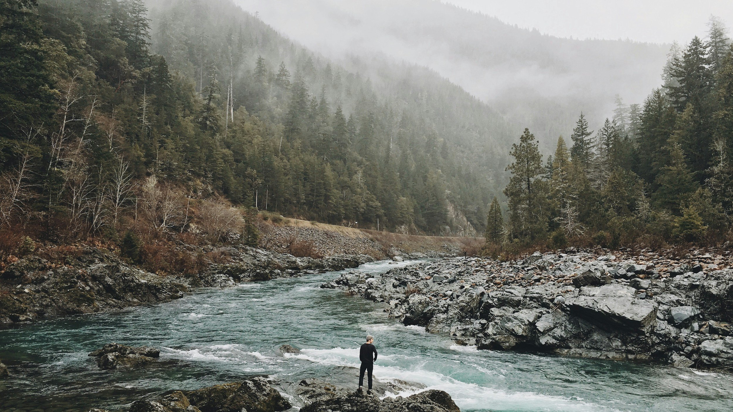 A person standing on rocks in a river with a forested mountain landscape and fog in the background.