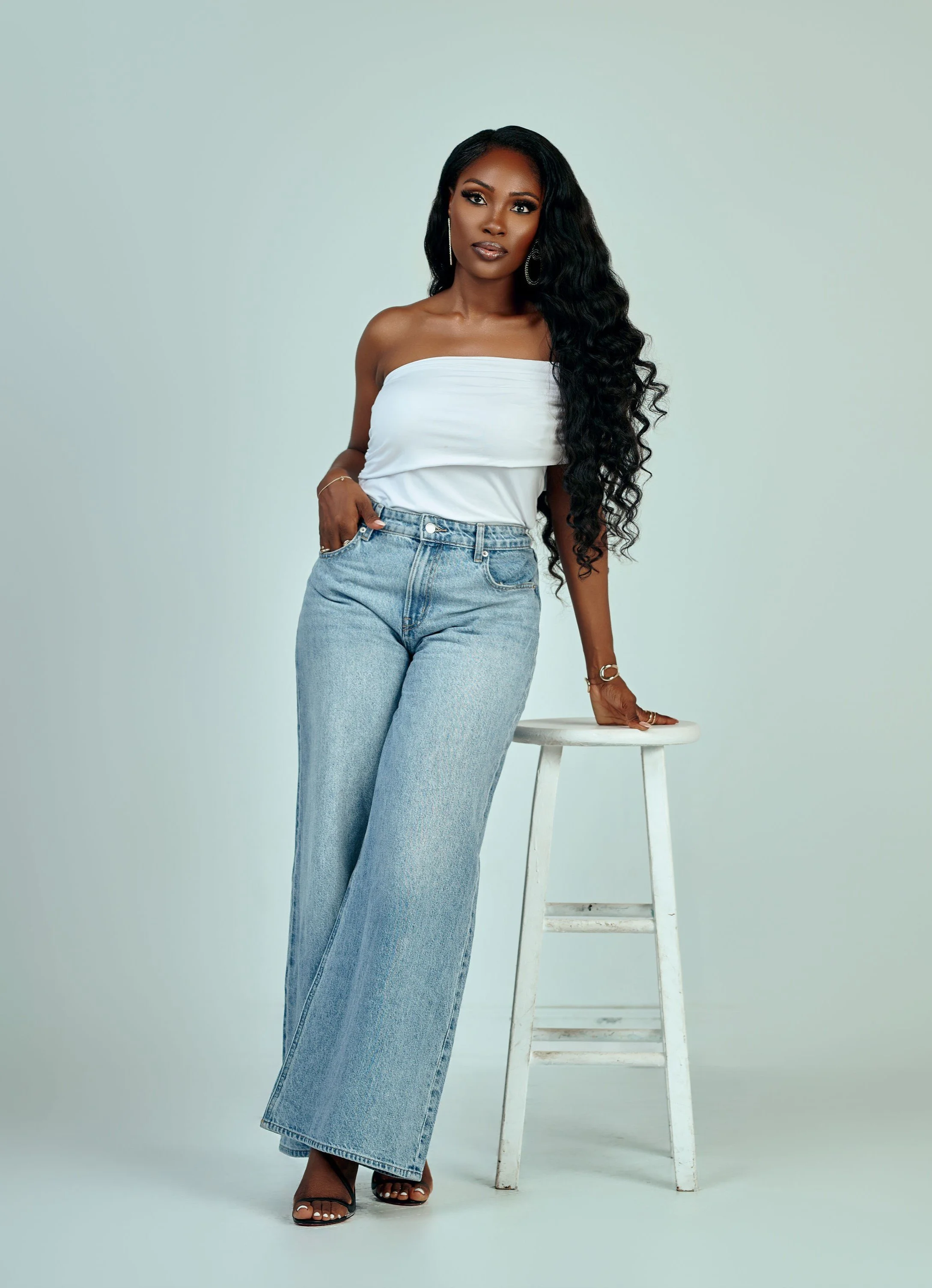 A woman with long, curly black hair and medium-dark skin tone standing next to a white stool against a plain light-colored background. She is wearing a strapless white top, high-waisted light blue jeans, and black heeled sandals.