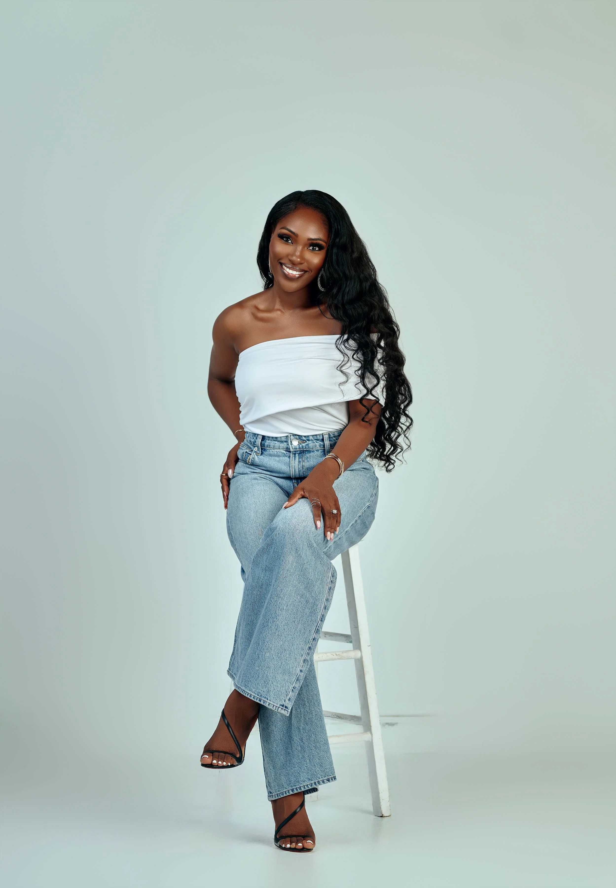 A woman with long, wavy black hair, smiling and sitting on a white stool against a plain light-colored background, wearing a white tube top, light blue jeans, and black high-heeled sandals.