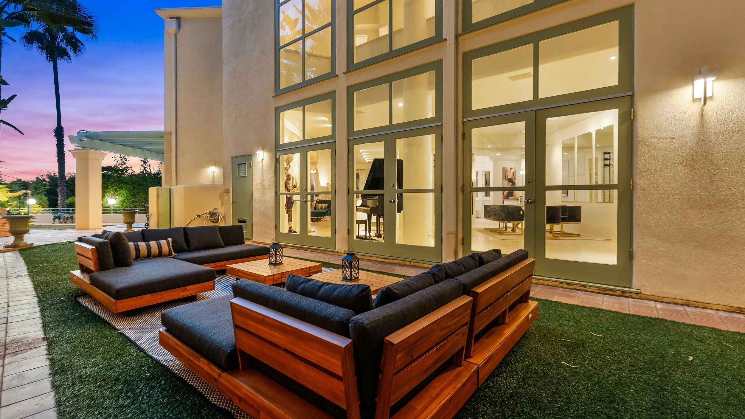 Outdoor patio area with black cushioned seating and wooden tables during sunset, adjacent to a large modern house with large glass windows revealing an interior with a grand piano, art pieces, and modern furniture.
