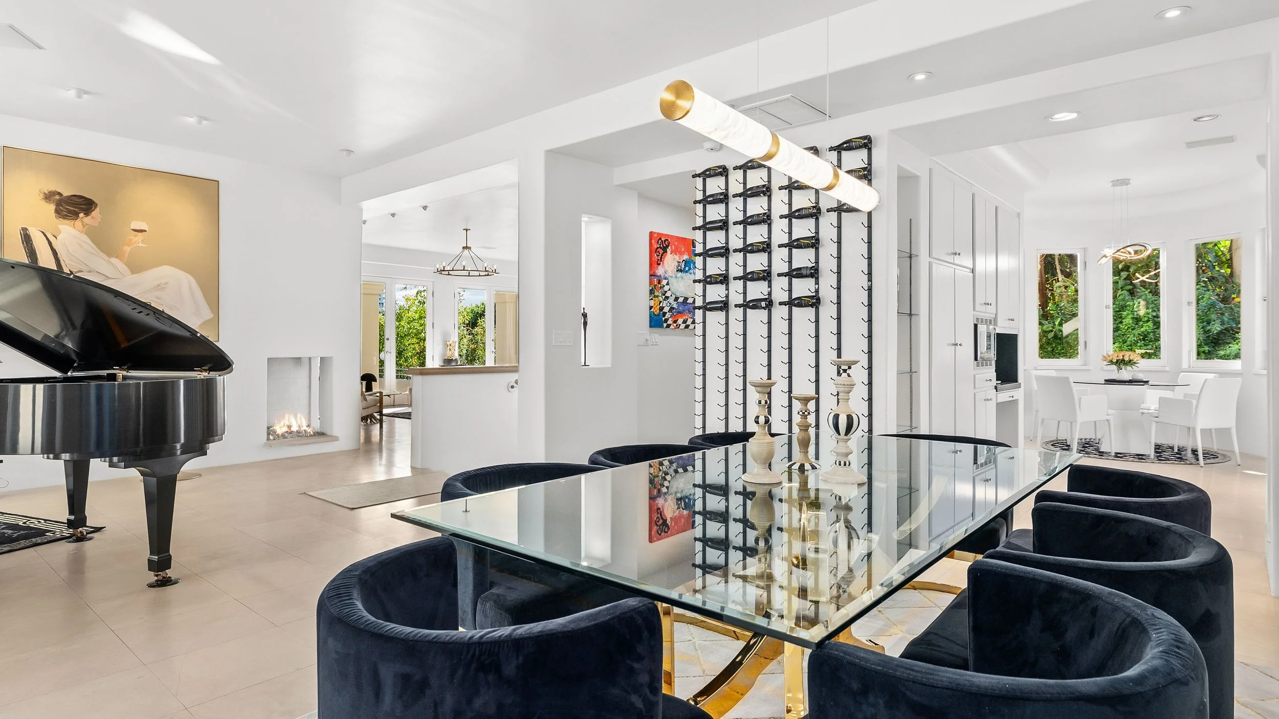 Modern living room with glass dining table, black velvet chairs, a grand piano, a fireplace, and art on white walls. Large windows with greenery outside.