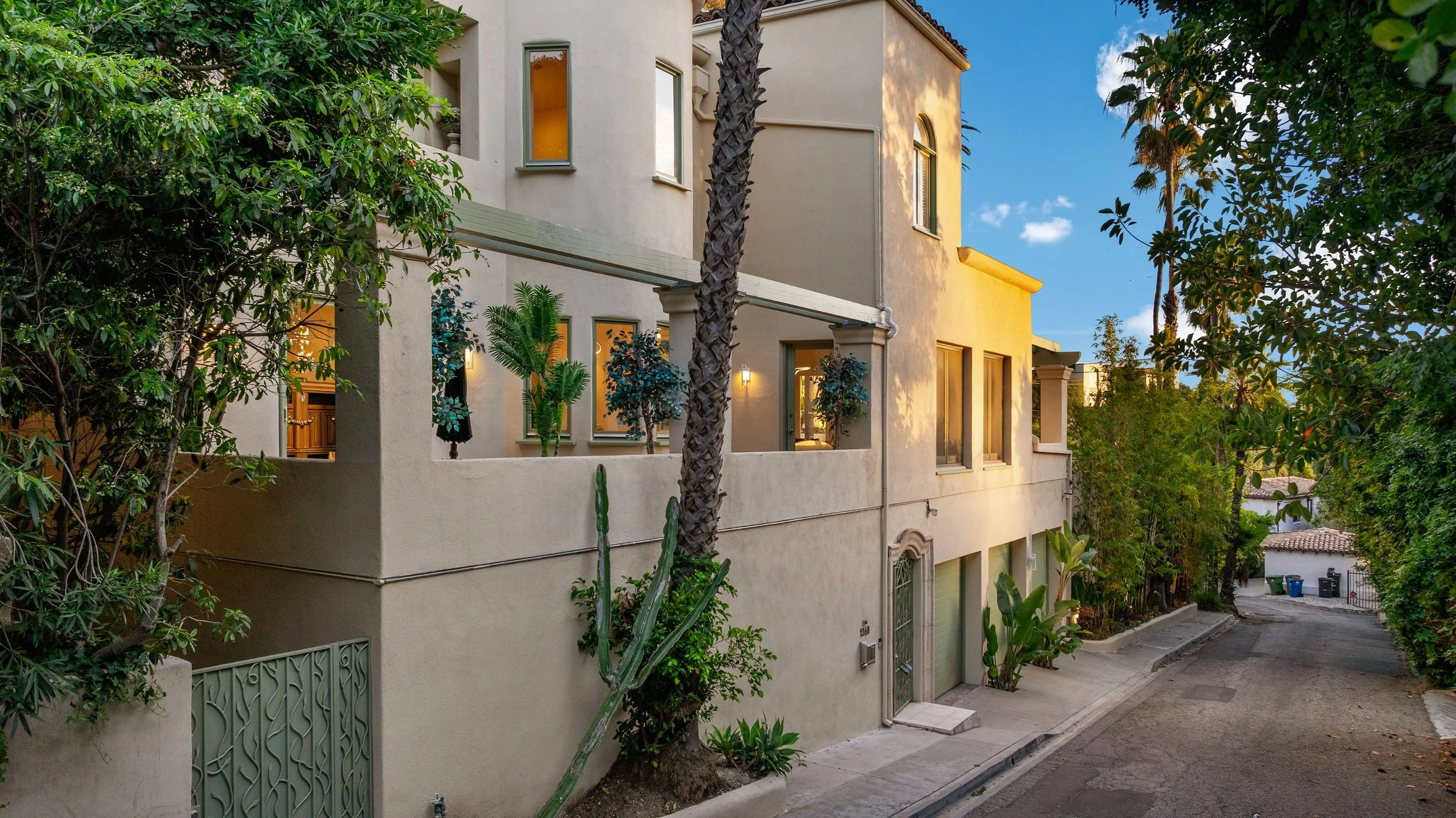 A multi-story residential building on a narrow street, with plants and trees including a tall palm tree and cactus, under a blue sky with a few clouds.