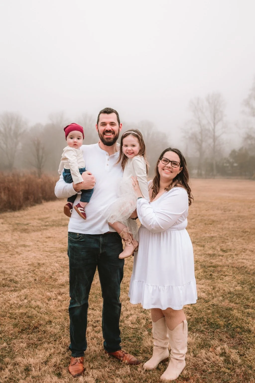 A happy family of four standing outdoors on a foggy day in a field. The father has a beard and is holding a young girl with a dress, and a young boy with a red cap is sitting on his arm. The mother, wearing glasses, is standing next to them with her arm around the girl. All are smiling.