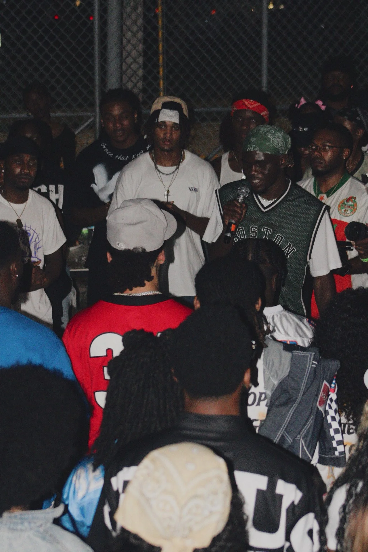 A group of young people gathered outdoors at night, with one man speaking into a microphone while others watch and listen.