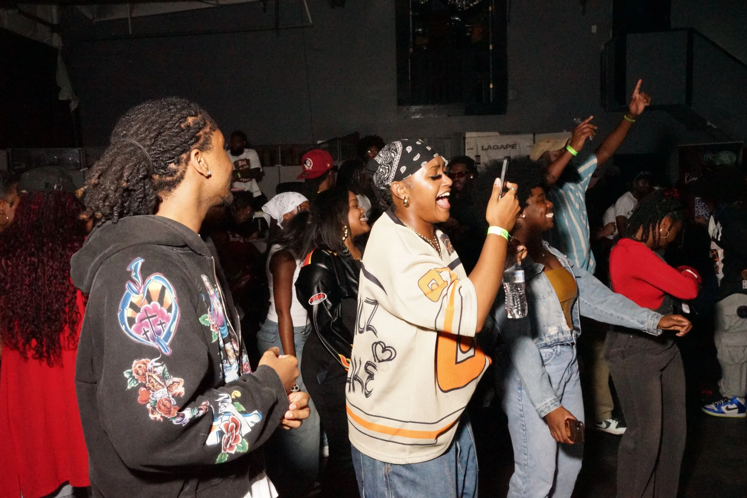 People enjoying a concert or event, dancing and singing along, with some taking pictures, in a dark indoor venue.