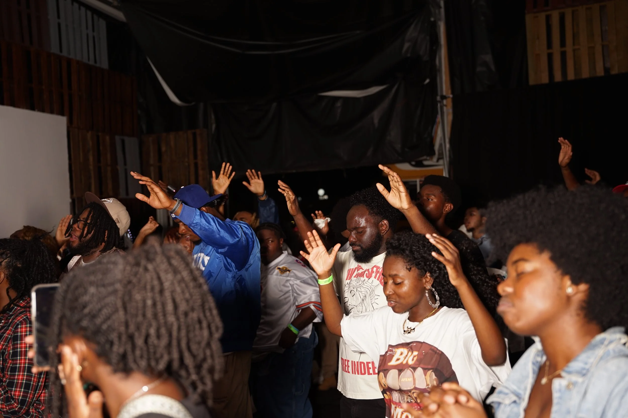 A group of people, mostly young, with their eyes closed and hands raised, participating in a worship or prayer session in a dimly lit indoor venue.