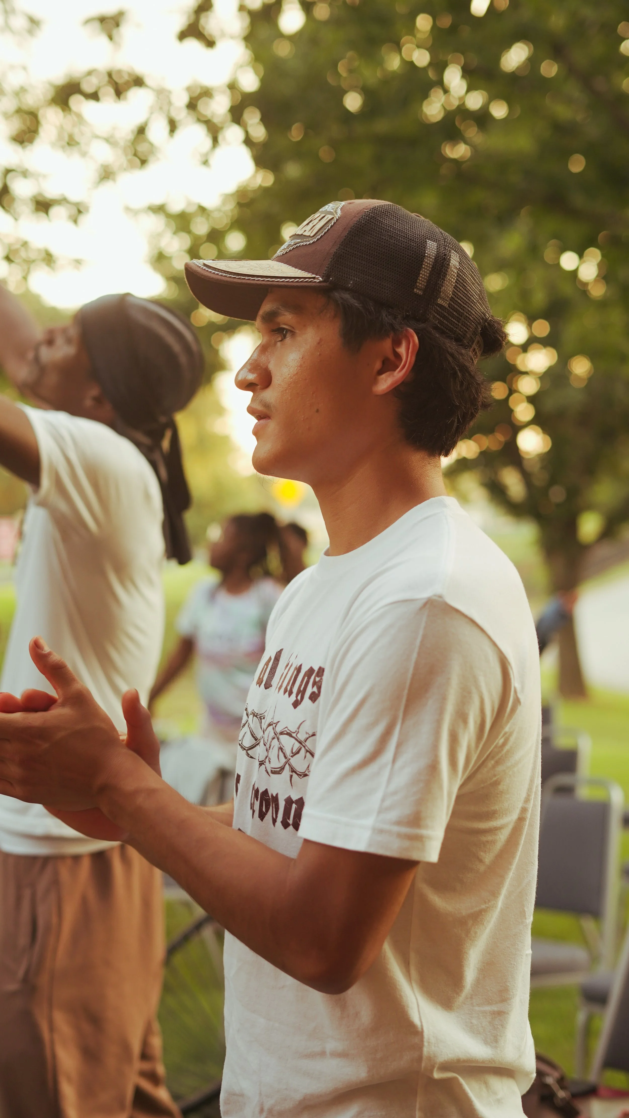 A young man wearing a trucker cap and a white T-shirt stands outdoors during daytime, with a blurred background of trees and other people.