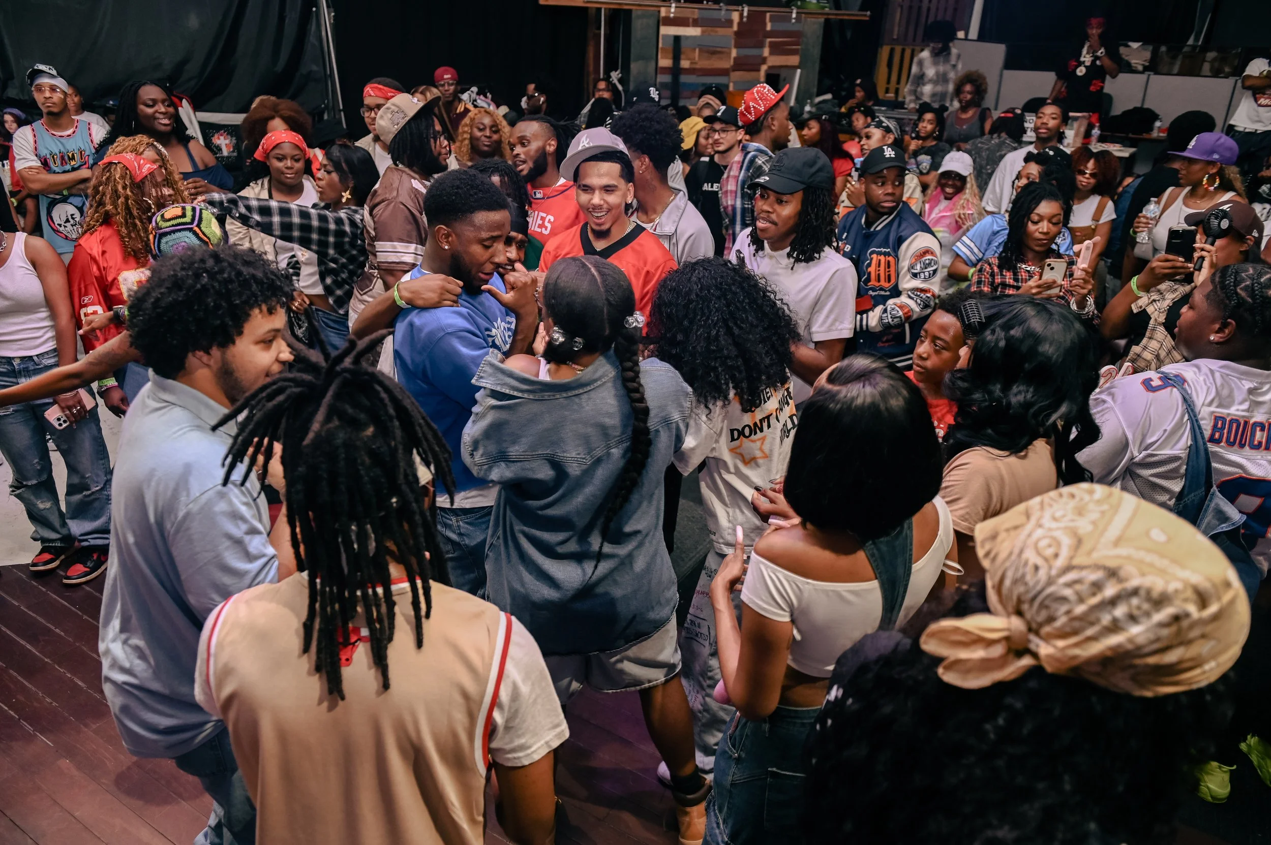 A large group of people gathered indoors at a social event or party, many wearing casual and colorful clothing, some taking pictures, smiling, and engaging with each other.