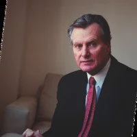 Man in a suit sitting on a beige sofa in a room with beige walls.