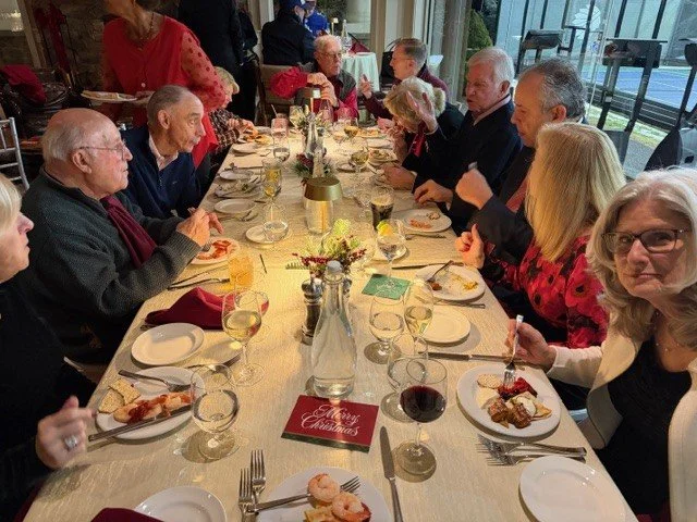People gathered at a long table for a holiday dinner, with plates of food, wine glasses, and a centerpiece with flowers.