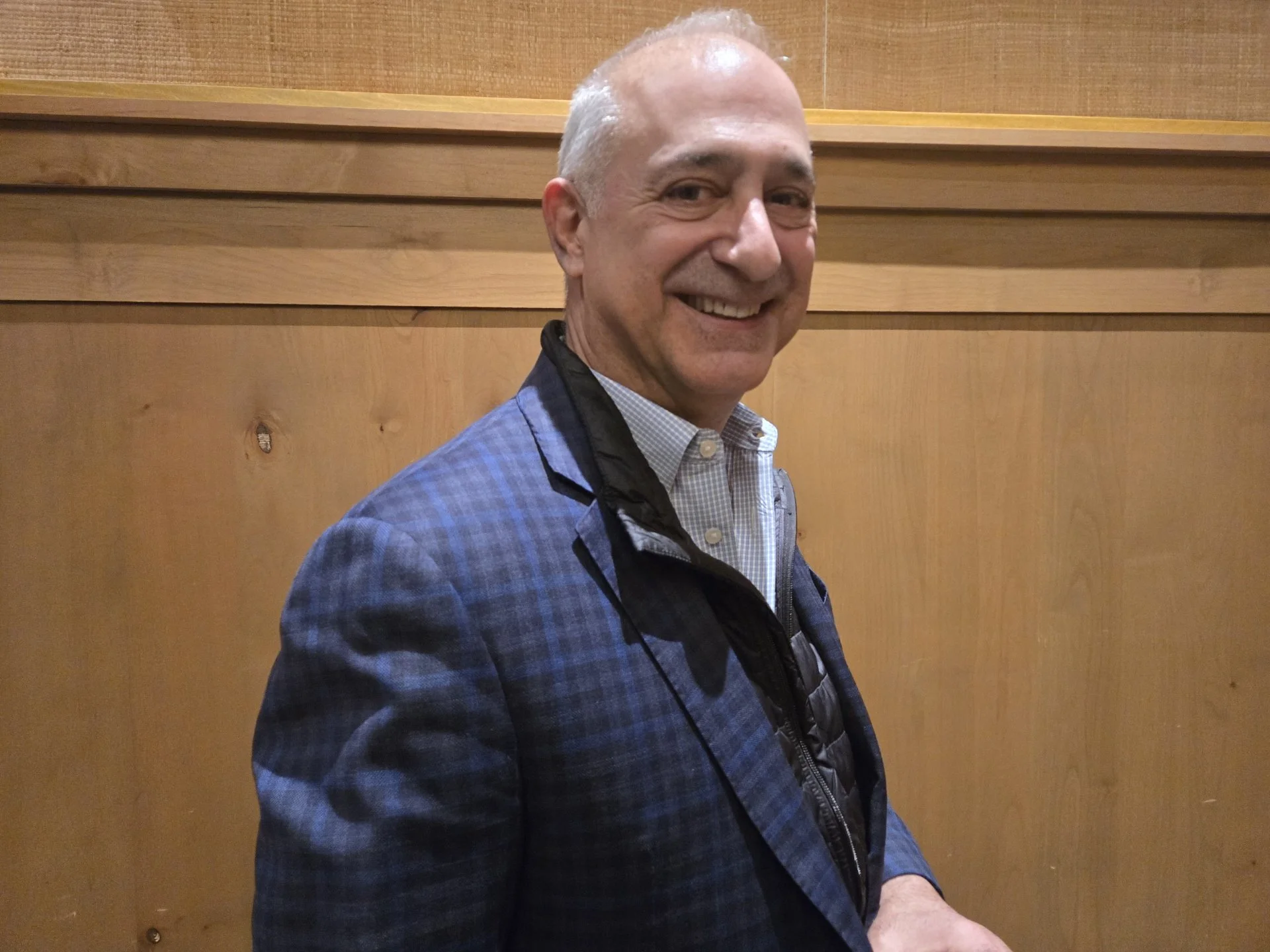 A smiling bald man wearing a patterned blue blazer and a light-colored collared shirt standing in front of a wooden-paneled wall.
